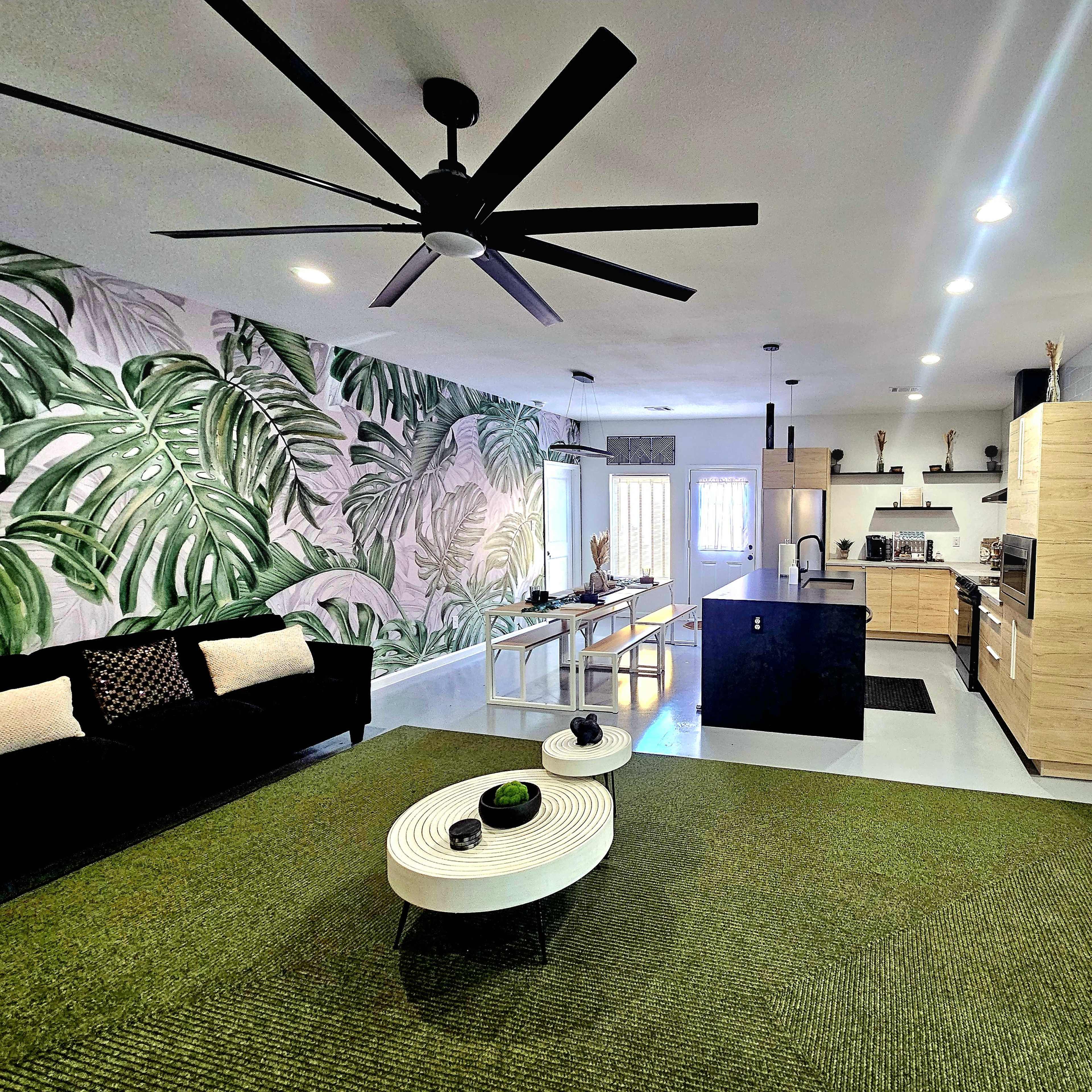 A spacious living area featuring a dark sofa, a round coffee table, a large green area rug, and a kitchen with light-colored cabinetry and a tropical leaf mural on the wall.