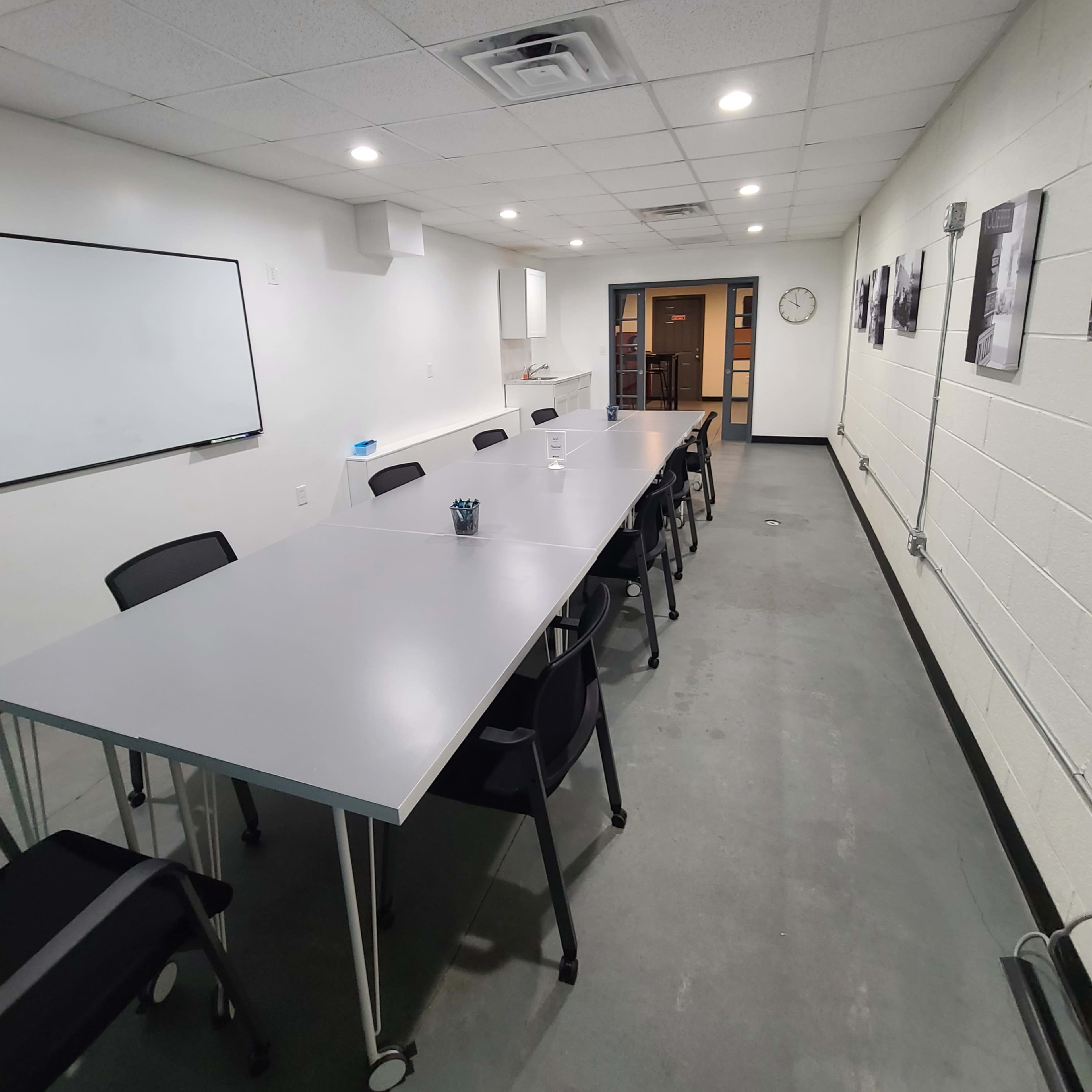 A long, rectangular conference room with a gray table, black chairs, a whiteboard, and a clock on the wall.