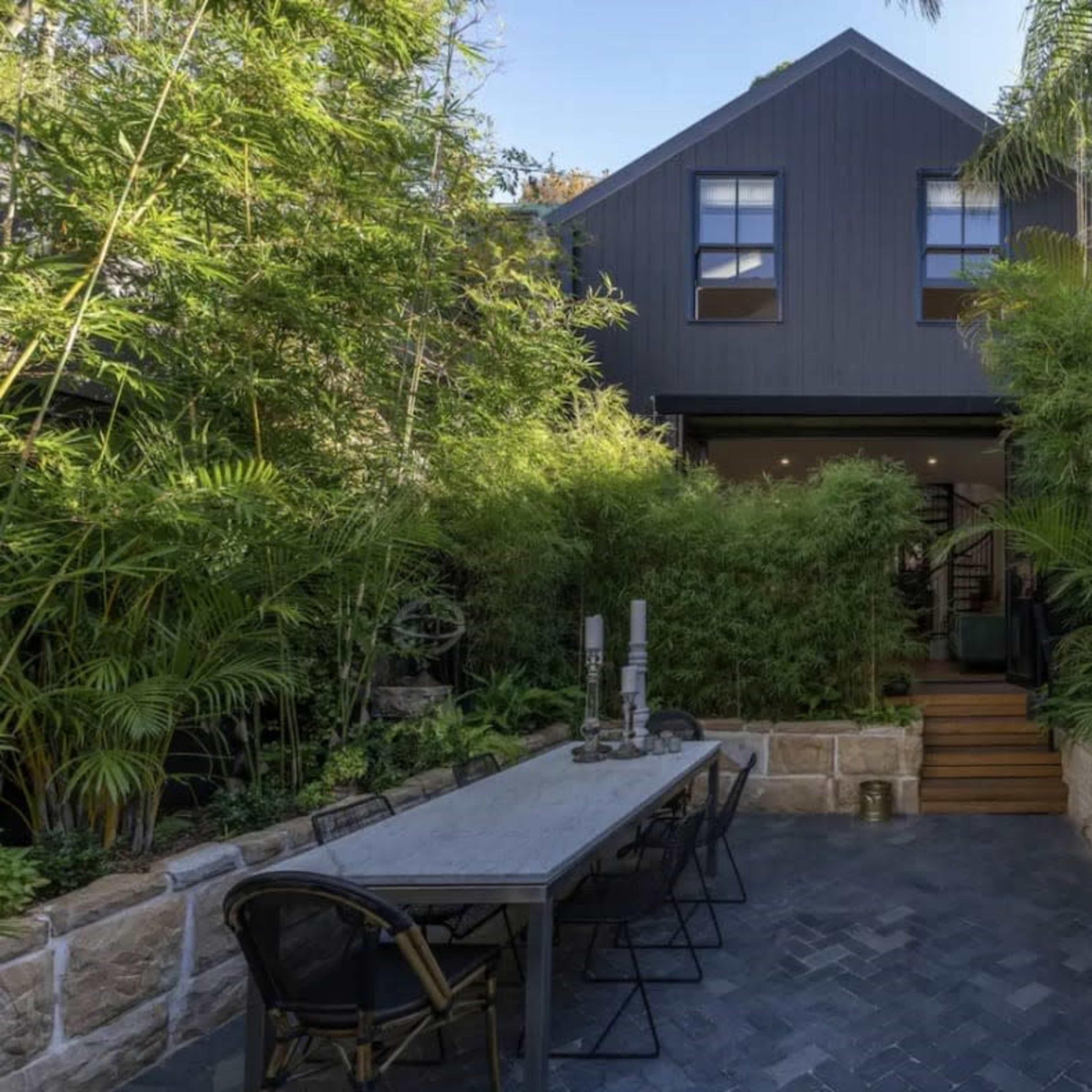 A black house surrounded by dense greenery, with a stone patio and a large dining table set in front.