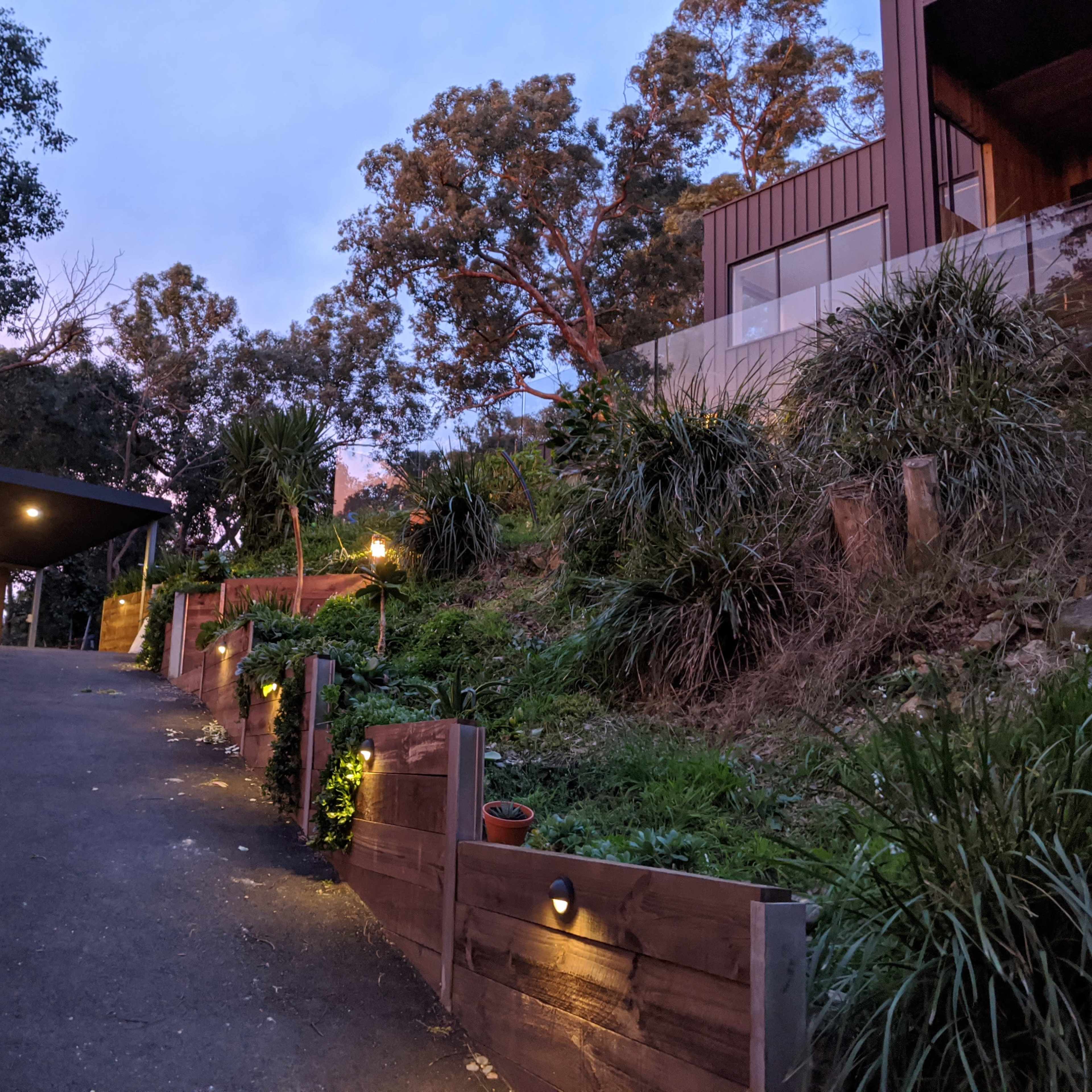 A driveway with a carport leads to a modern house, surrounded by landscaped greenery and illuminated by garden lights at dusk.
