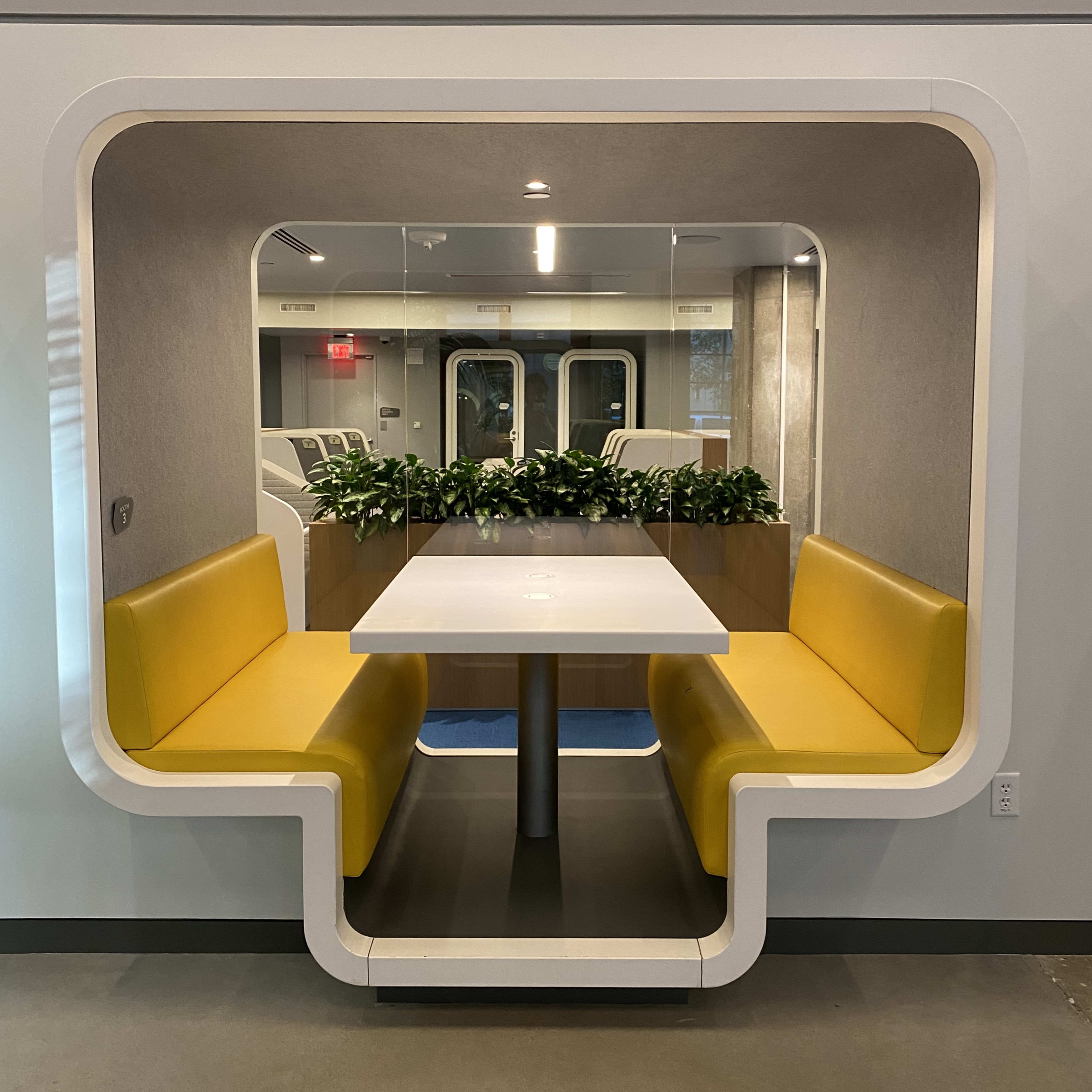 A modern booth with yellow seating and a white table, framed by a square arch and adorned with greenery.