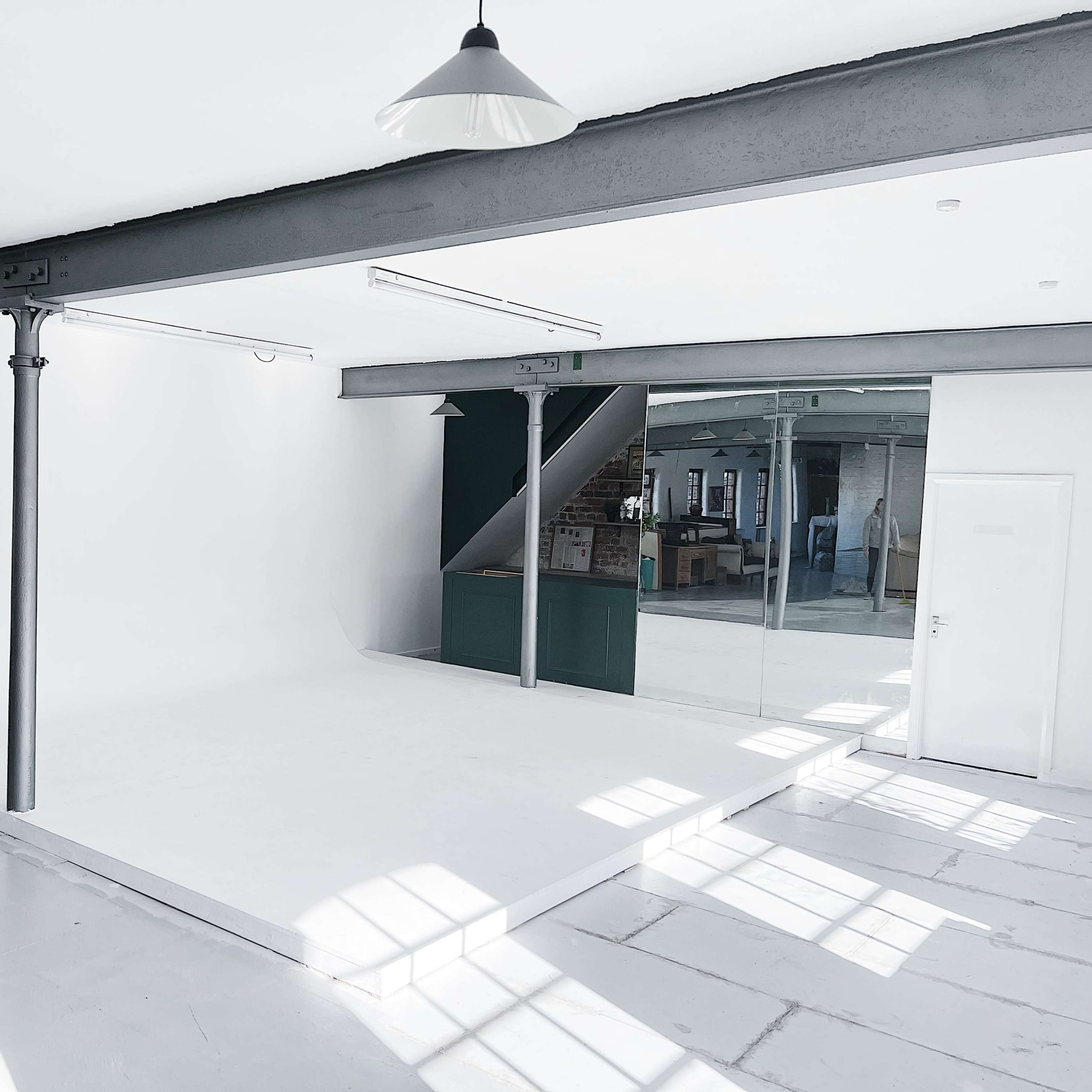 A minimalist studio space featuring a white backdrop, large windows allowing natural light, and industrial-style beams and columns.