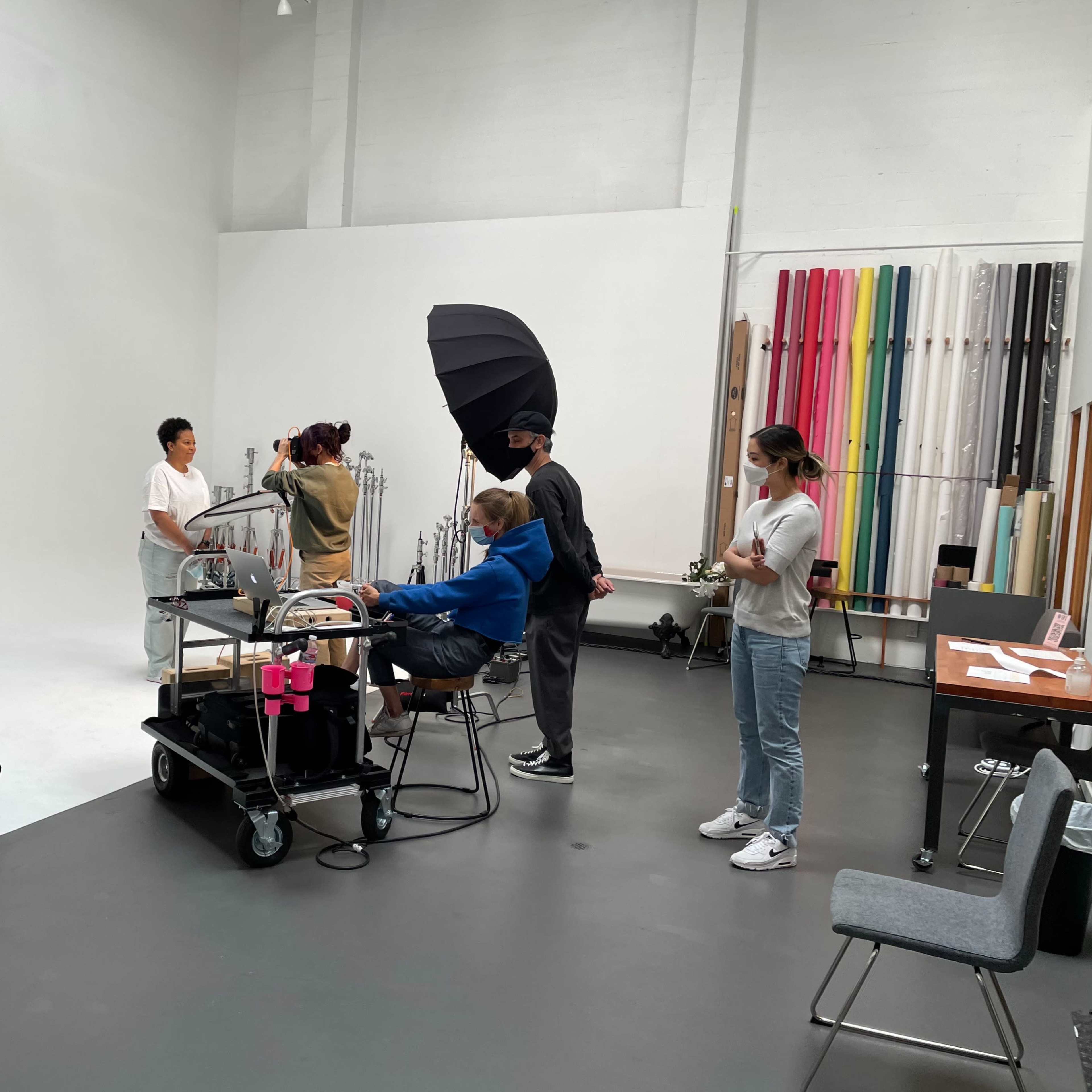 A group of five people are working in a studio space equipped with background rolls and lighting setups, while one person is seated at a mobile workstation.