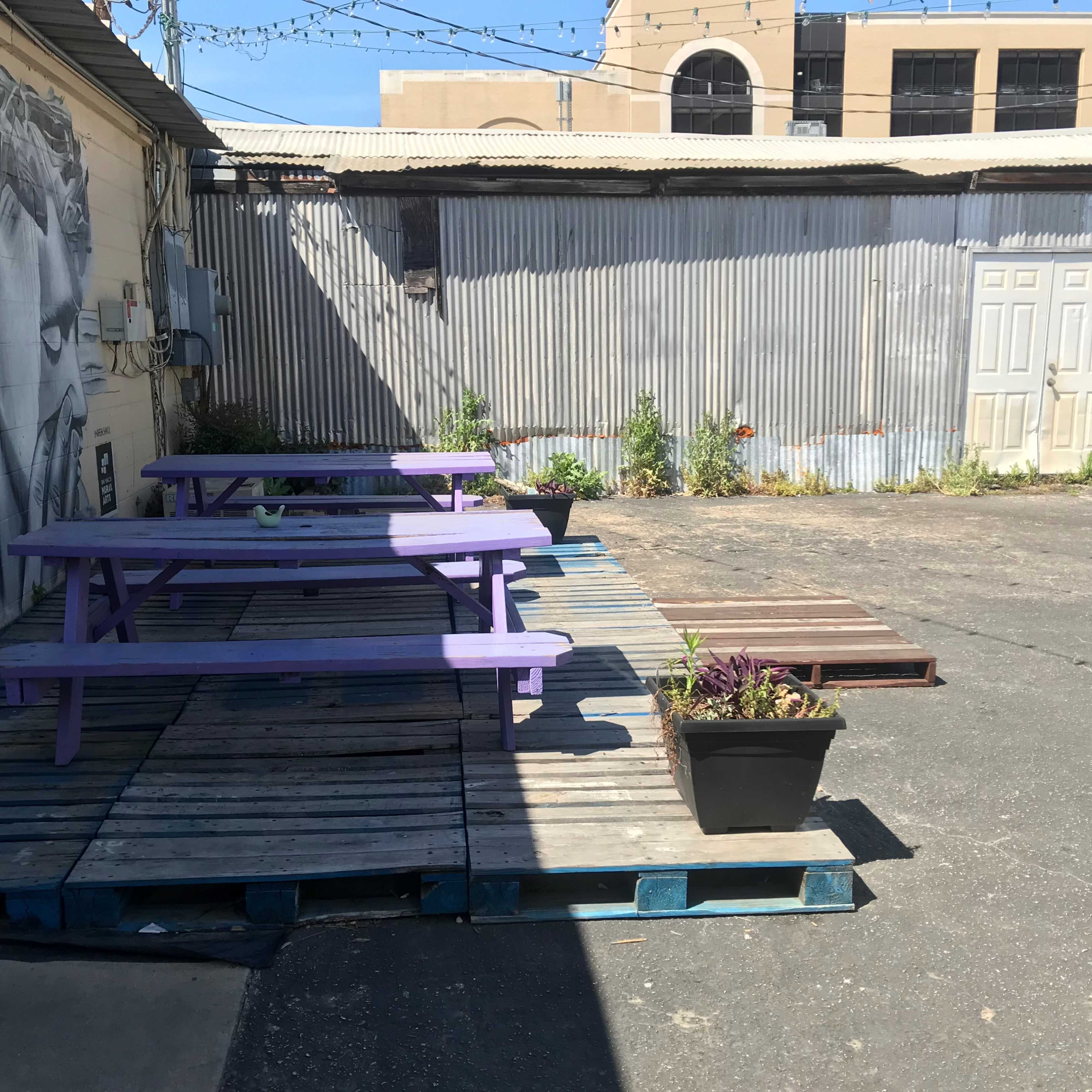 A fenced outdoor area with purple picnic tables on wooden pallets and a corrugated metal backdrop.