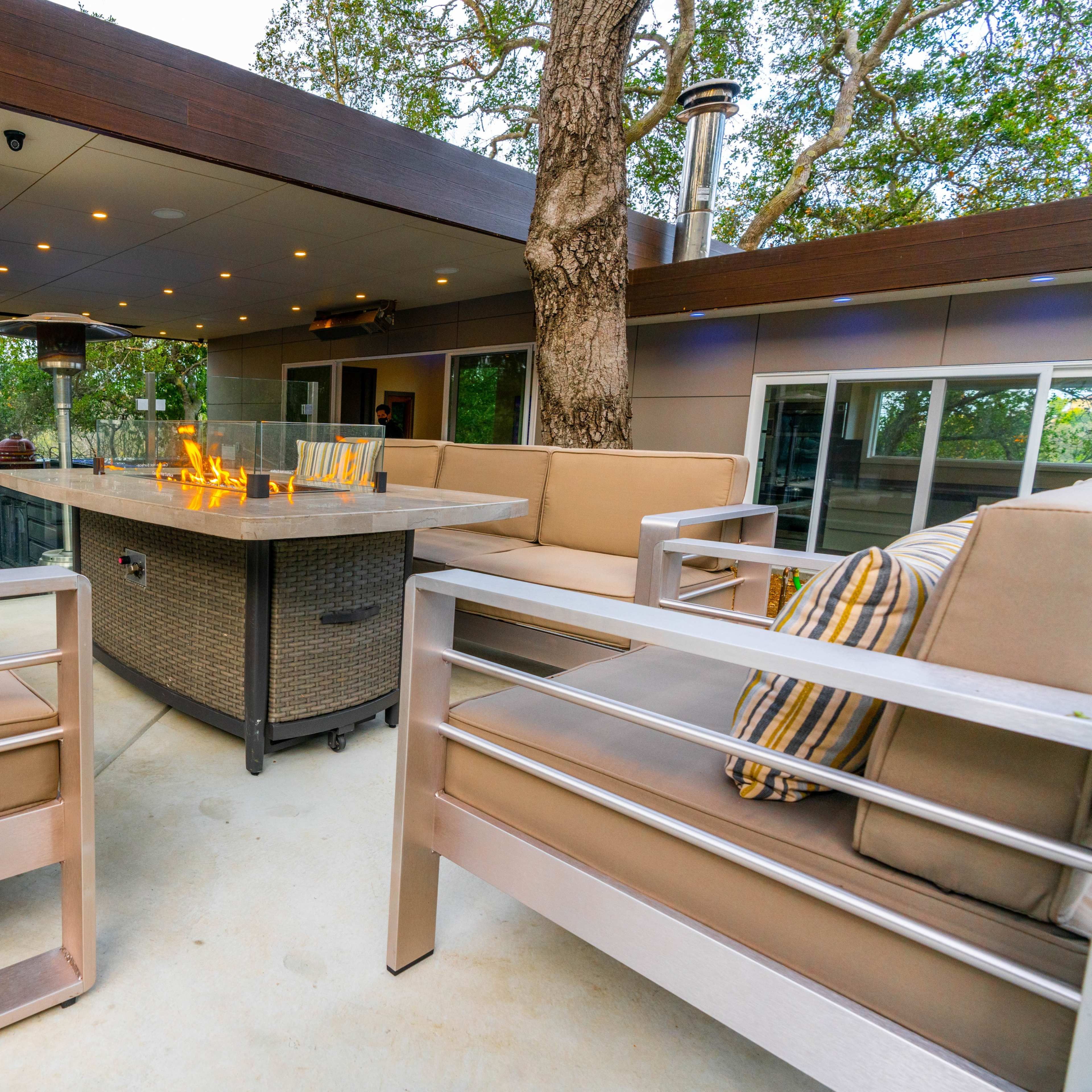 A modern outdoor lounge area with a central fire table surrounded by seating and large windows set amidst trees.