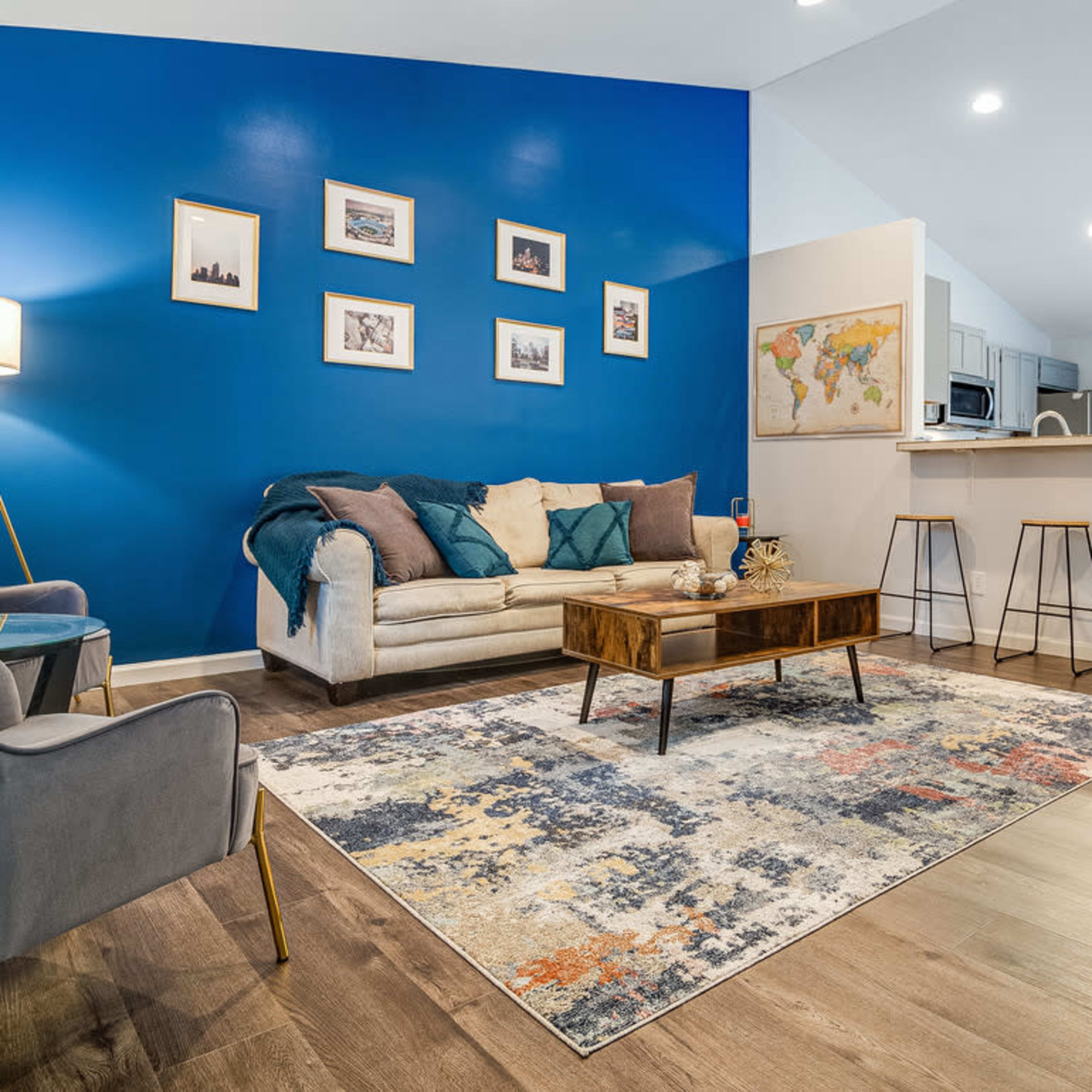 A modern living room with blue accent walls, a light-colored sofa, a wooden coffee table, and a dining area visible in the background.