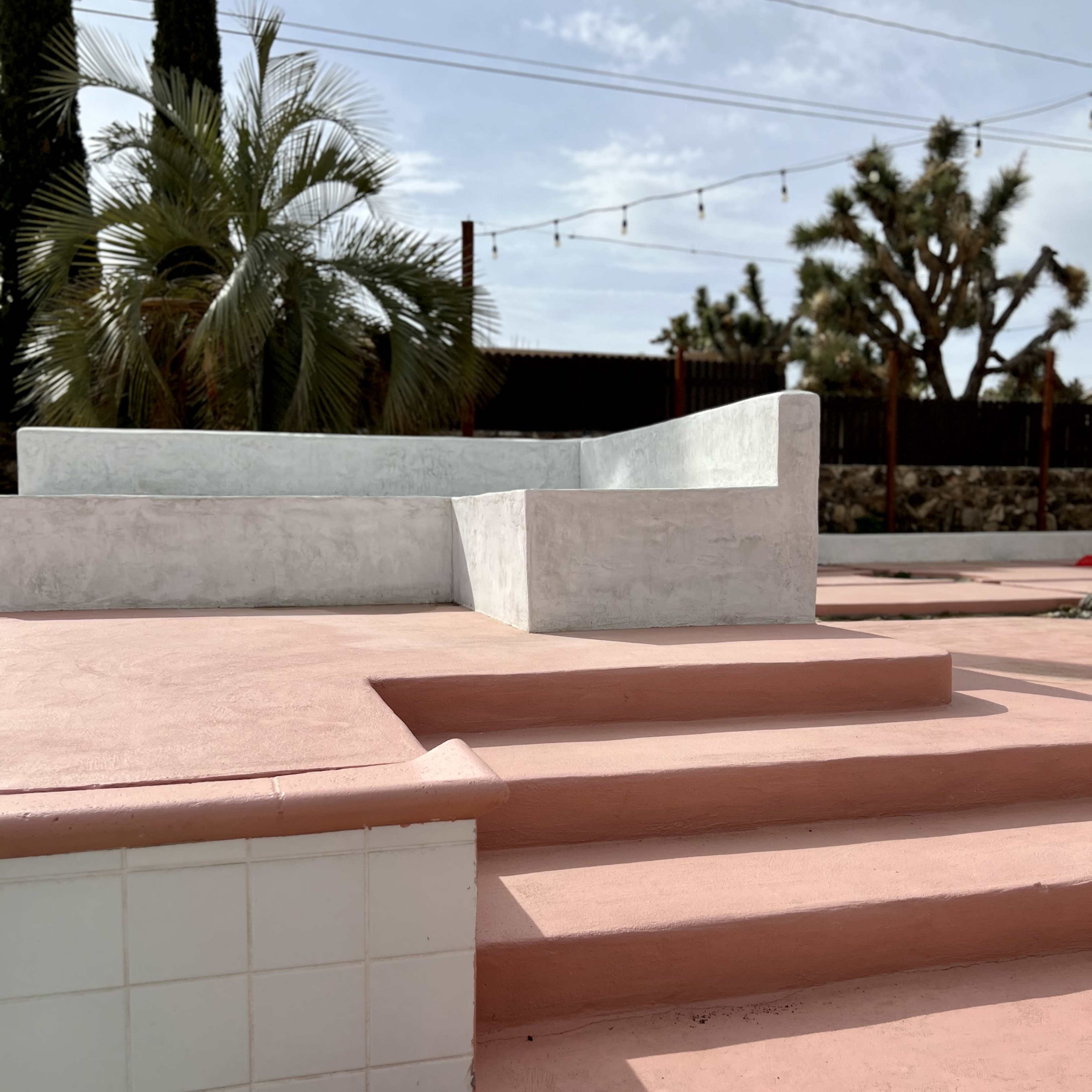 A pink patio area with a concrete seating structure and steps leading up to it, surrounded by palm trees and other greenery.
