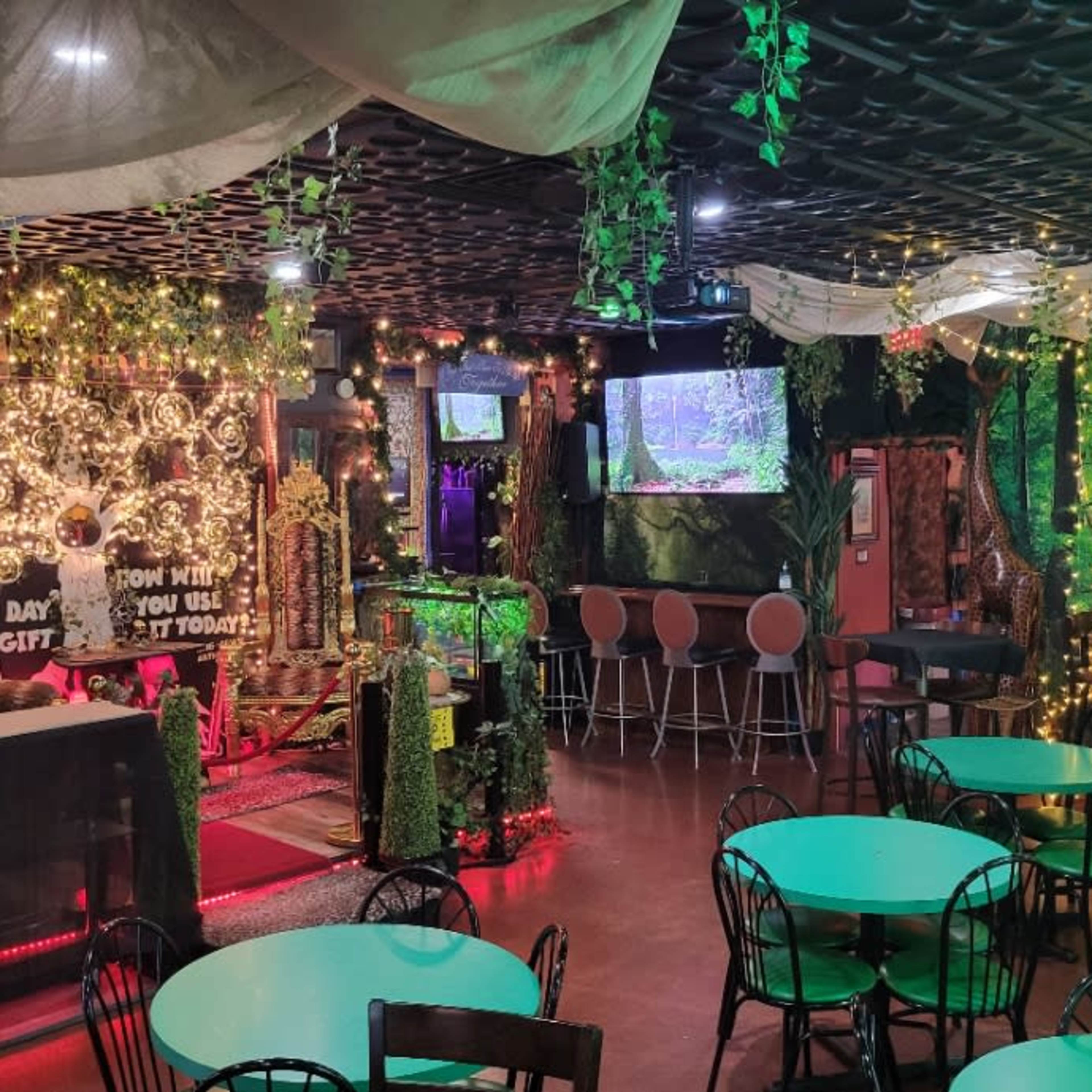 A vibrant, decorated restaurant interior with greenery, colorful lighting, and multiple screens displaying visuals.