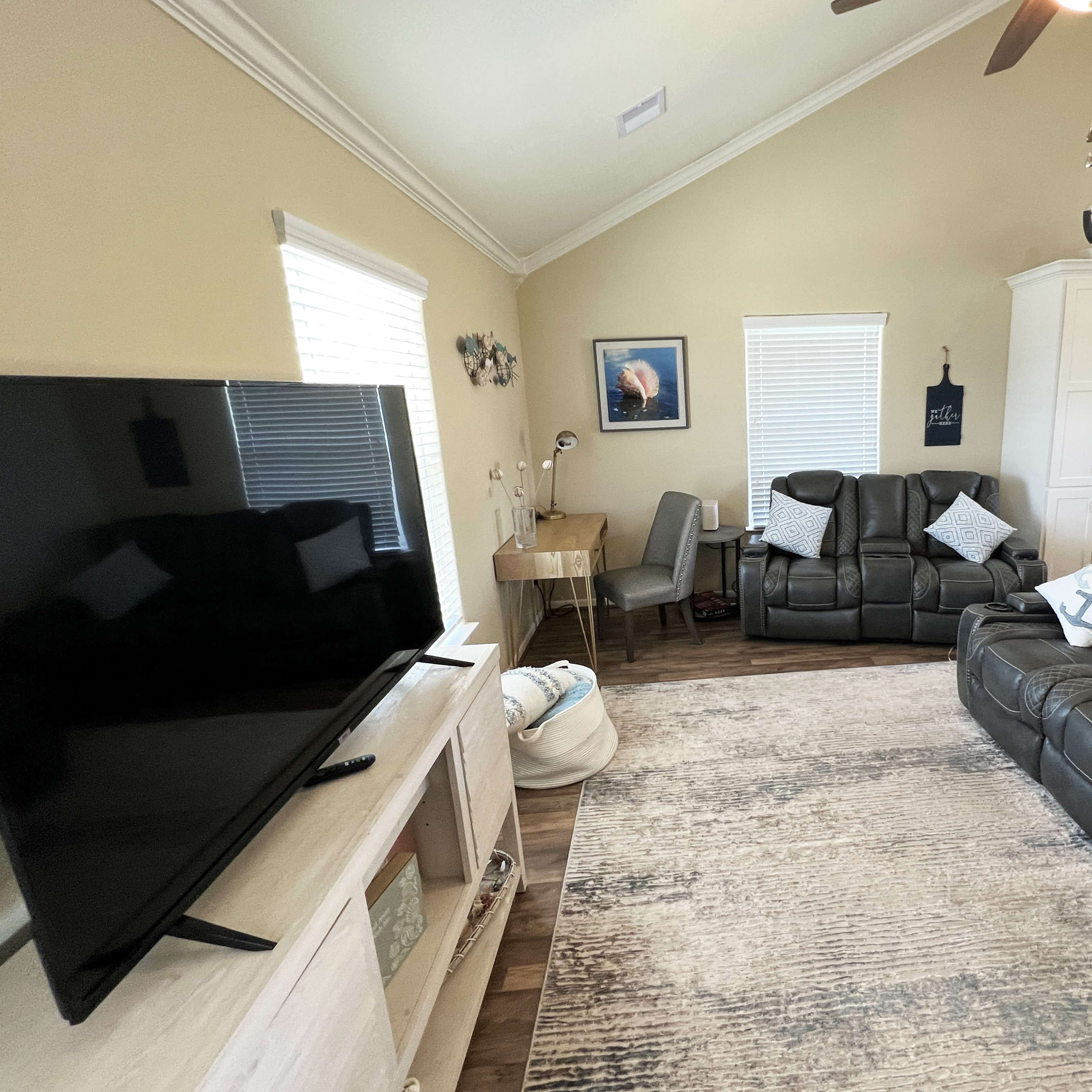 The living room features a large television, two reclining sofas, a desk chair, and a light-colored area rug under a vaulted ceiling.