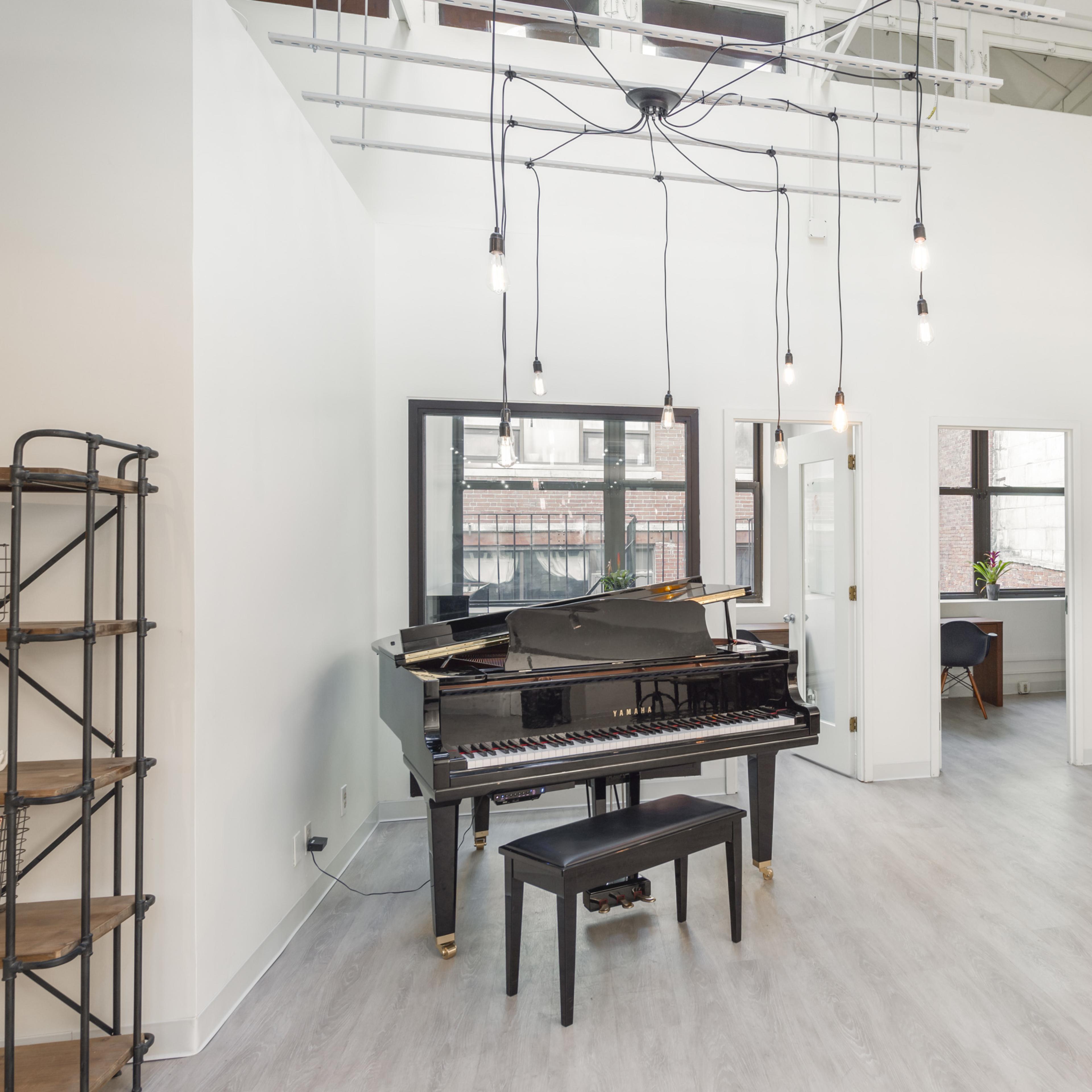 A bright room featuring a grand piano with a black stool, large windows, and modern lighting fixtures.