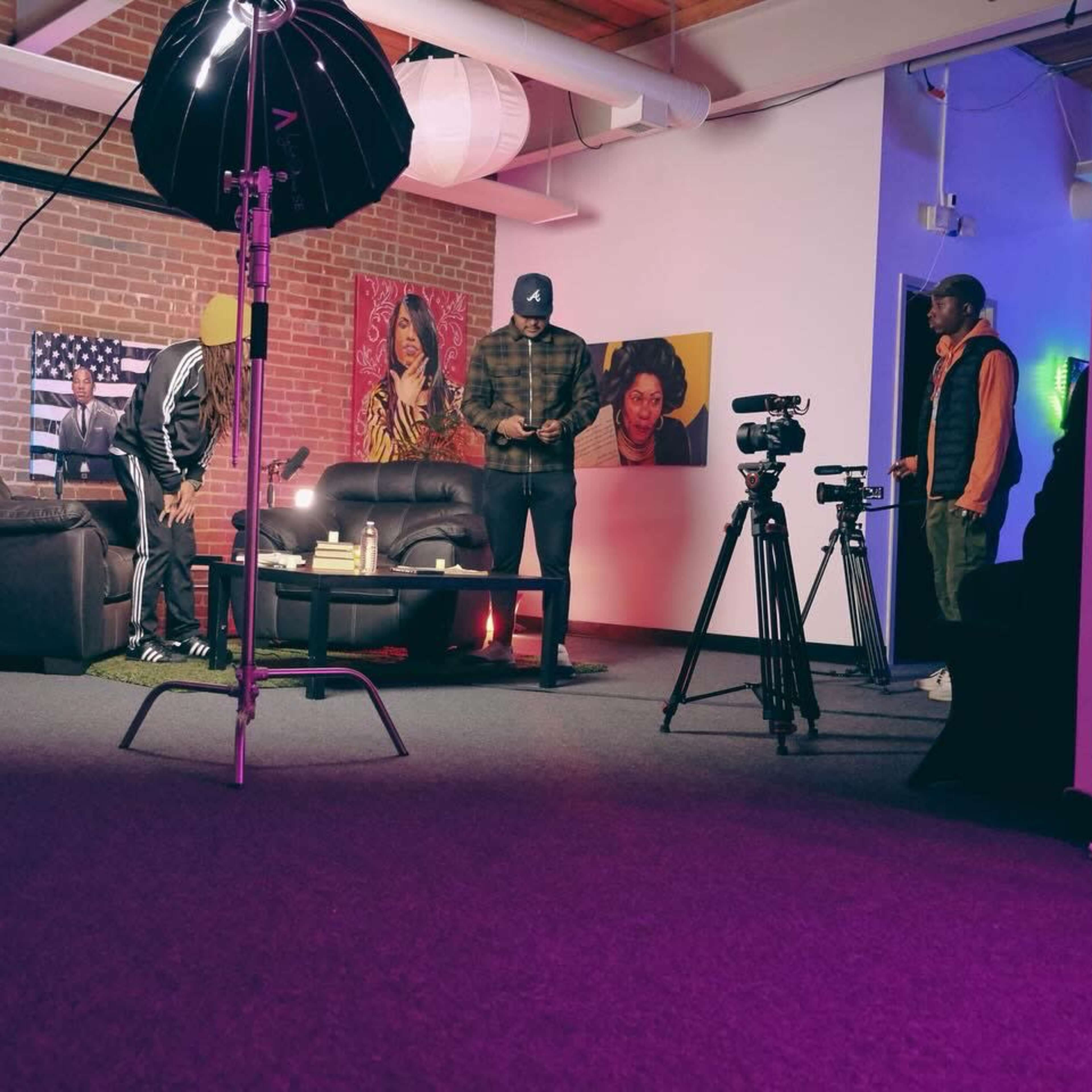 A group of four individuals prepares for a video shoot in a studio with colorful lighting, a black couch, and artwork on the walls.