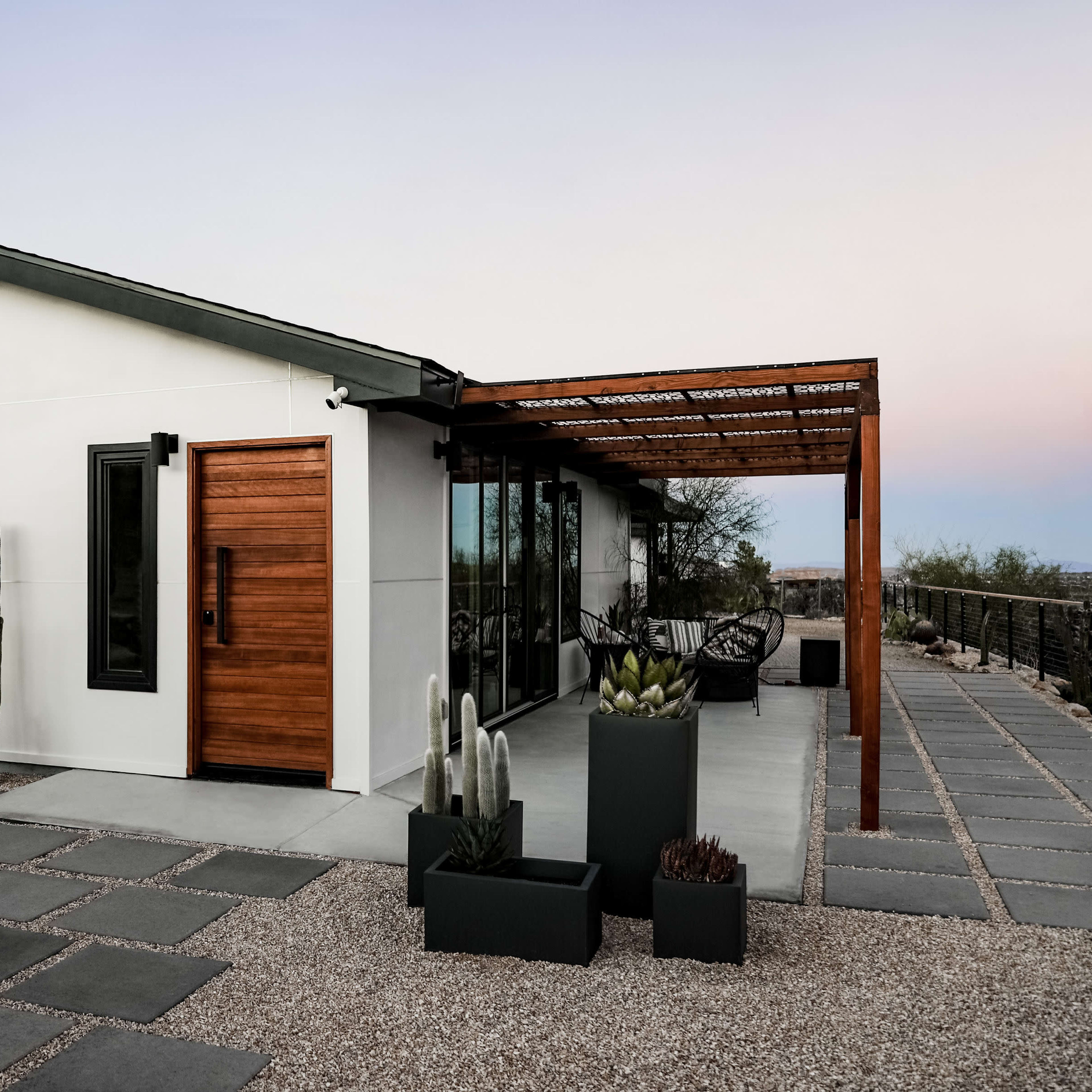 A modern house with a wooden front door and large windows is surrounded by gravel, cacti, and stone pathways.