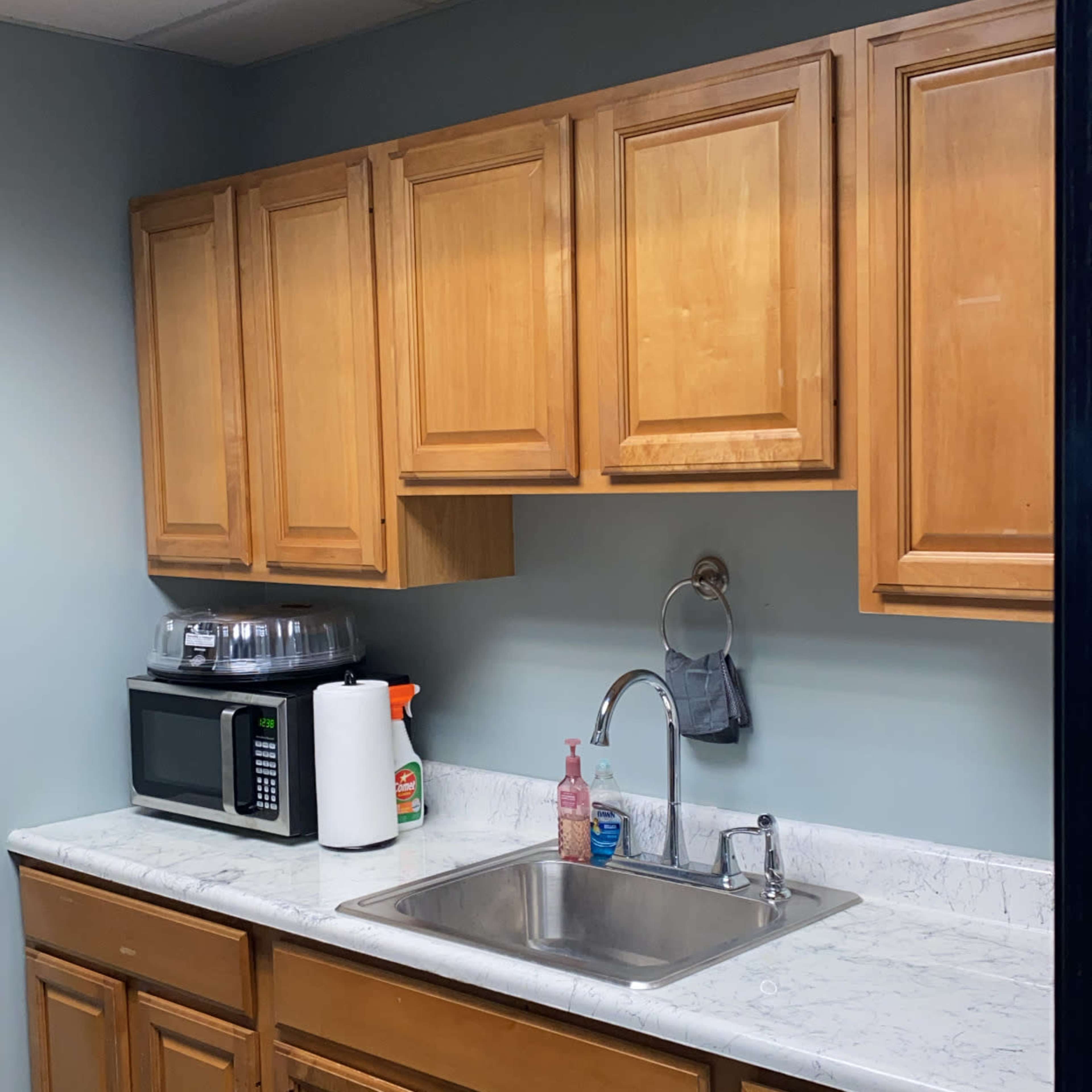 A kitchen area with wooden cabinets, a countertop with a sink, a microwave, and a soap dispenser on the side.