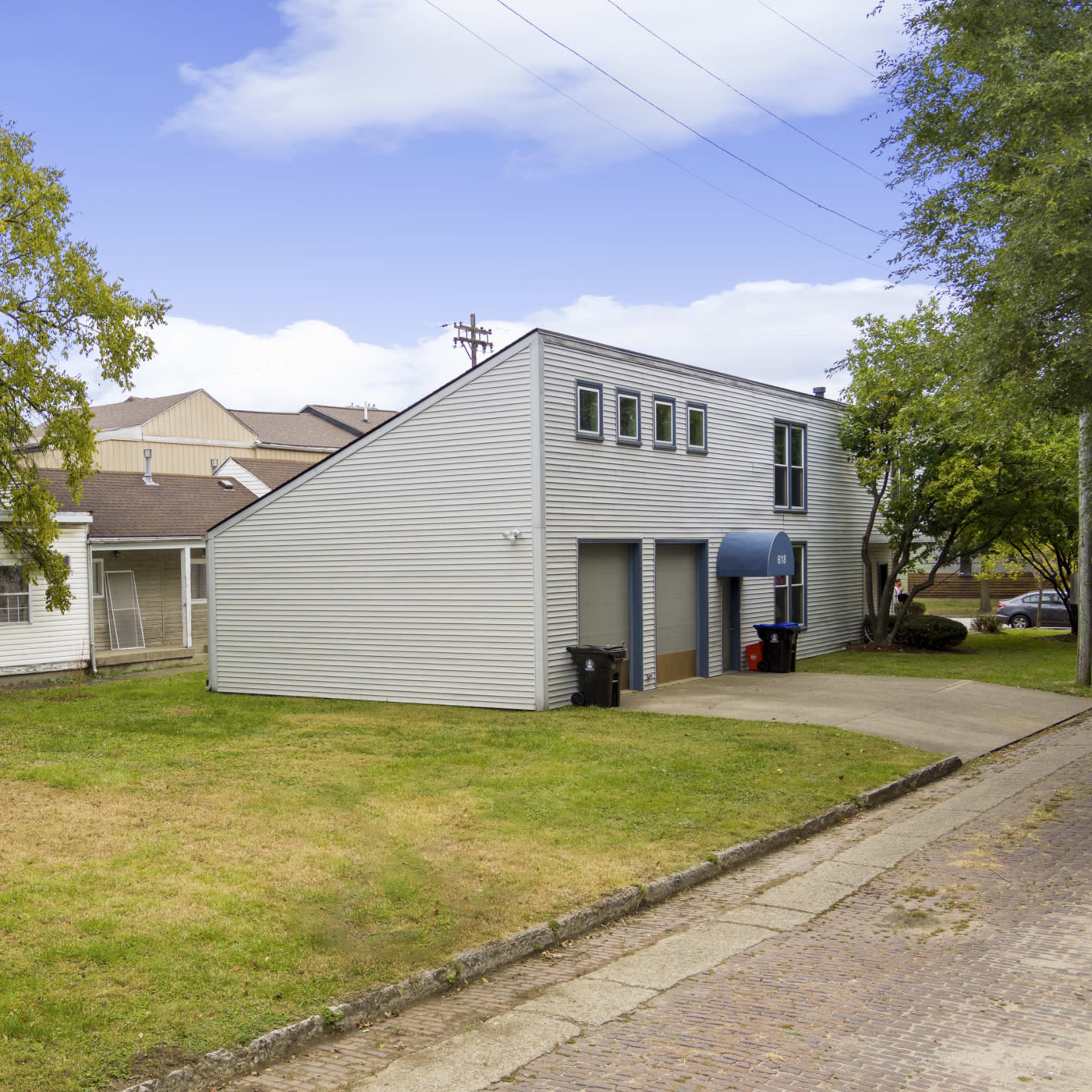Two buildings, one with a sloped roof and blue awning, situated on a grassy area beside a paved driveway.