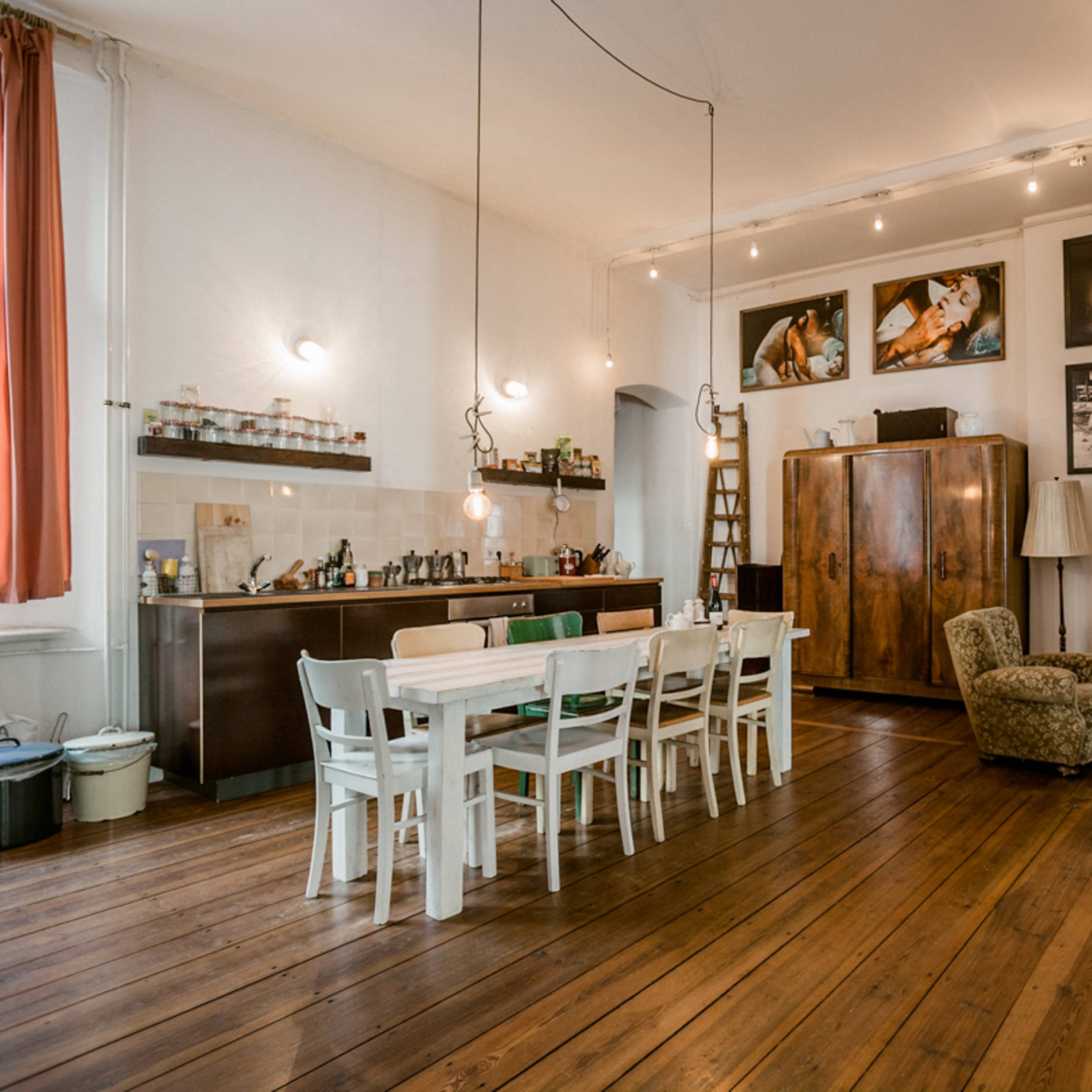 A spacious room with a kitchen area, a dining table, and a seating area, featuring wooden floors and various framed photographs on the walls.