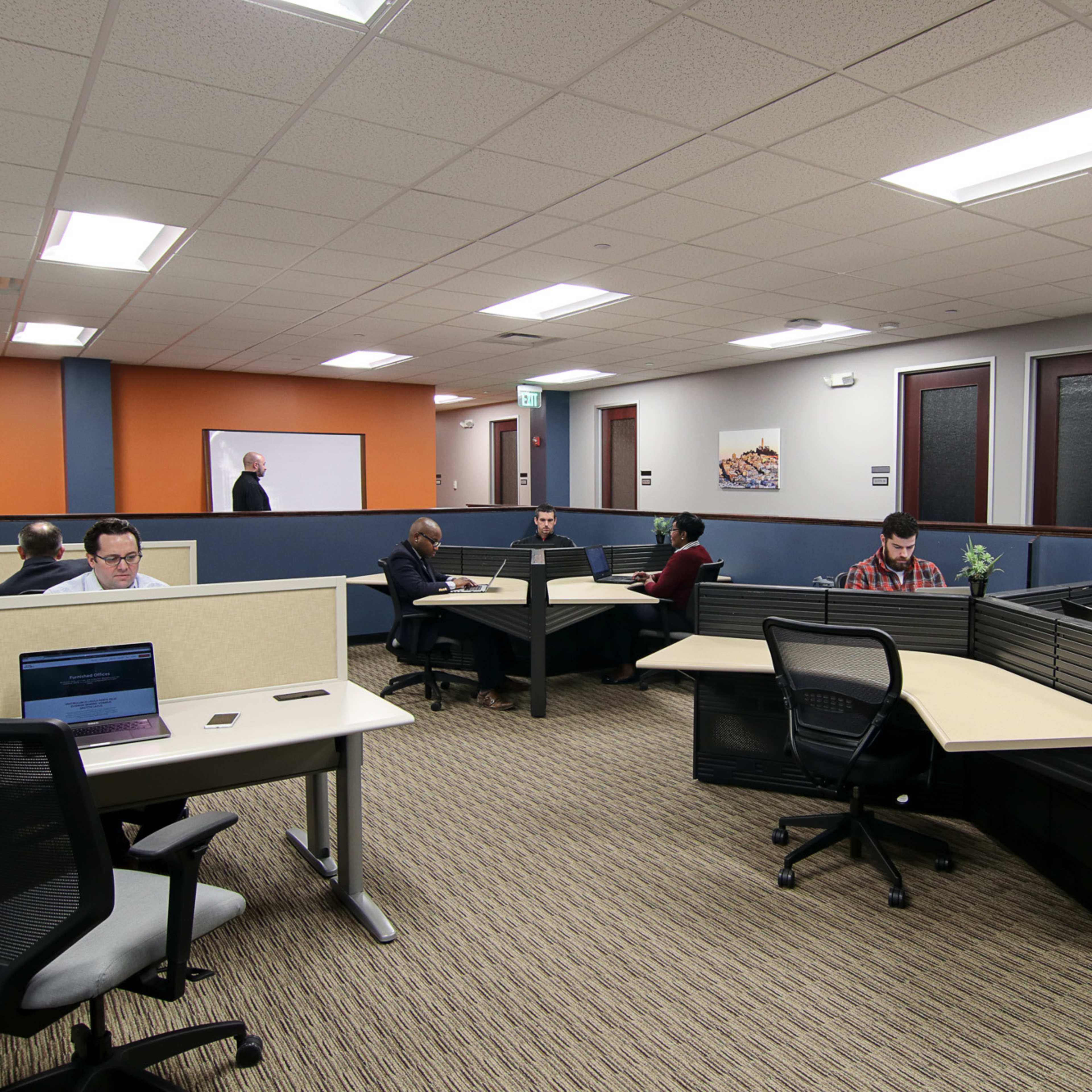 A modern office space with several workstations occupied by individuals using laptops in a well-lit environment.
