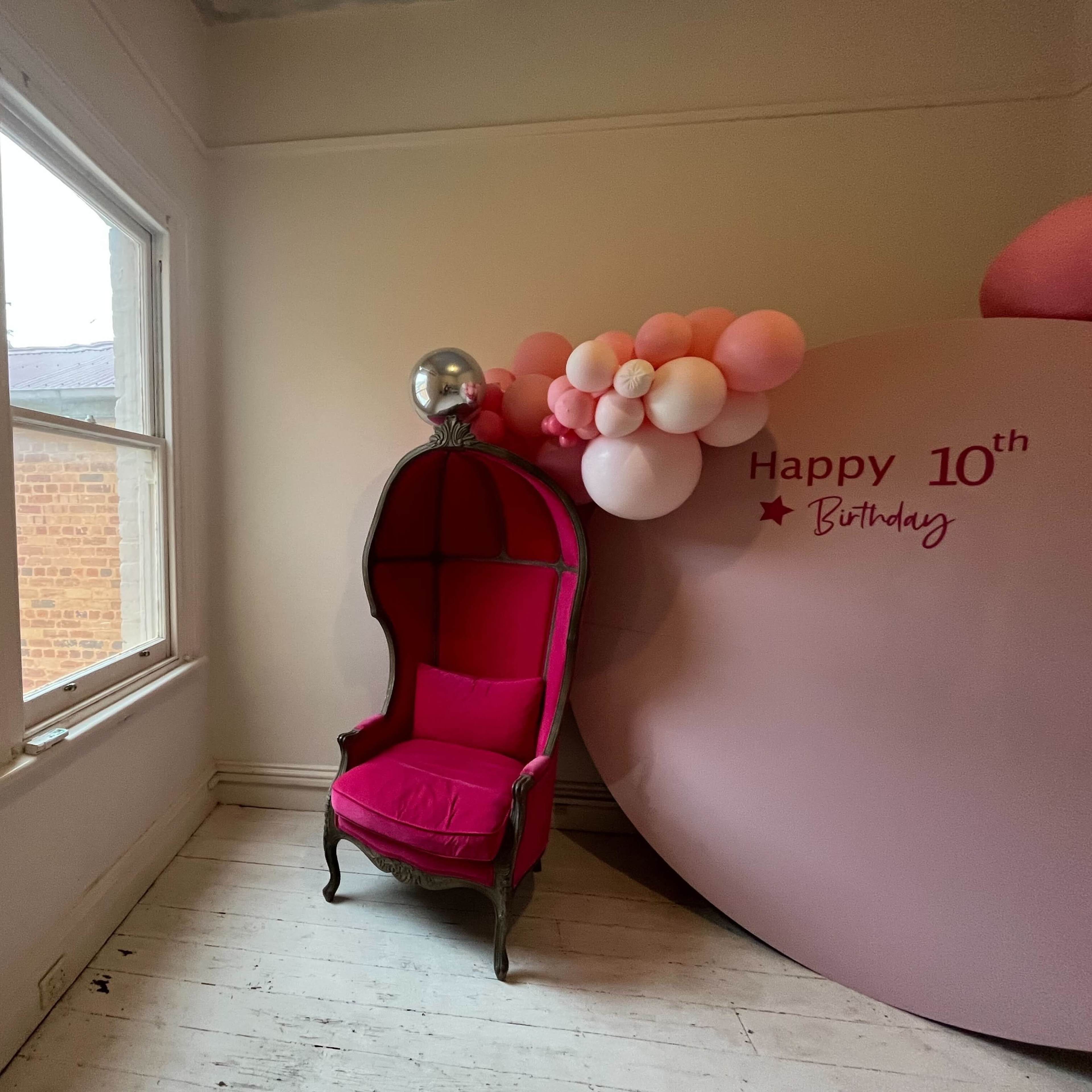 A pink chair in a room with a large round decoration that reads "Happy 10th Birthday" and a cluster of balloons nearby.