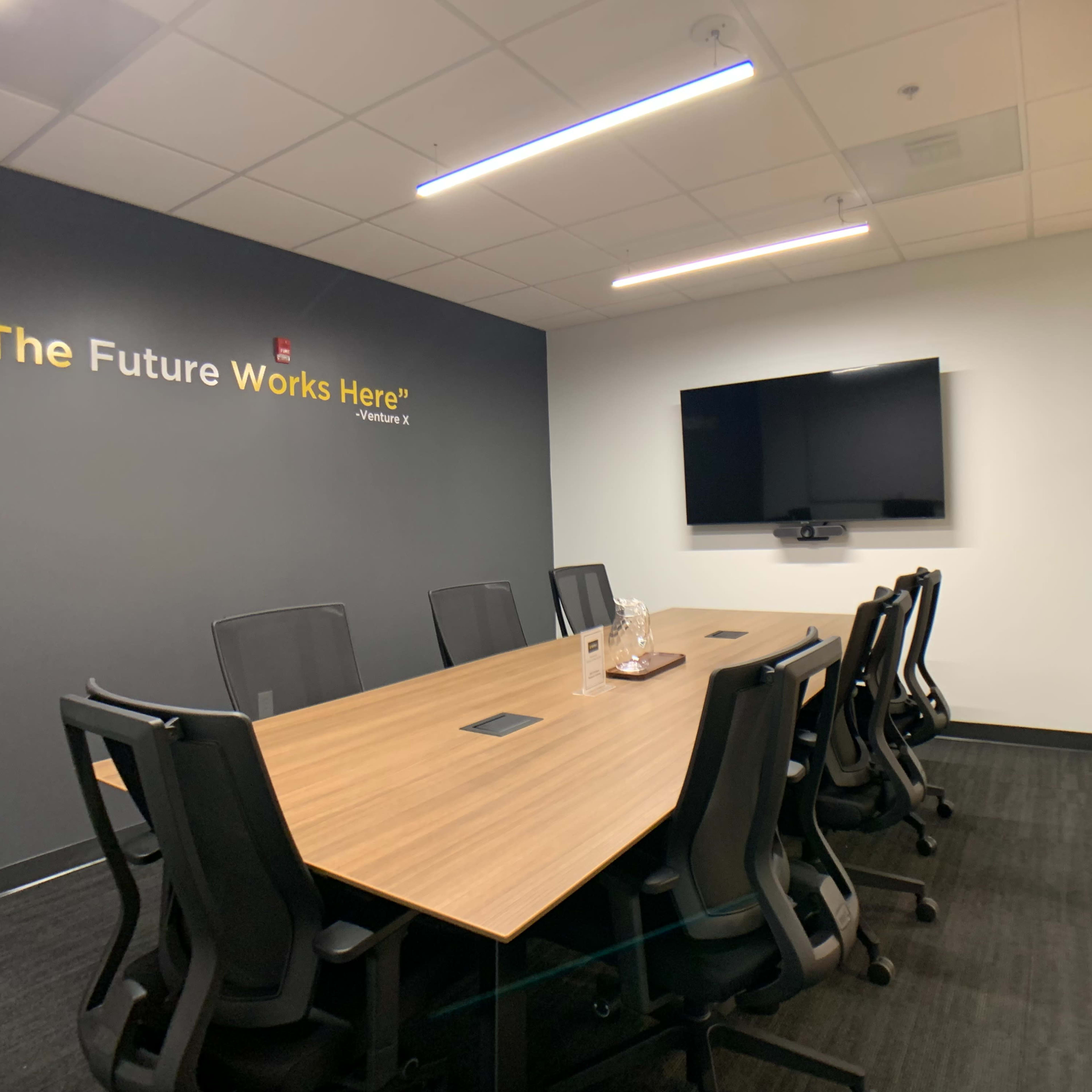 A modern conference room features a large wooden table surrounded by black chairs, a mounted television, and a wall with the phrase "The Future Works Here" above it.