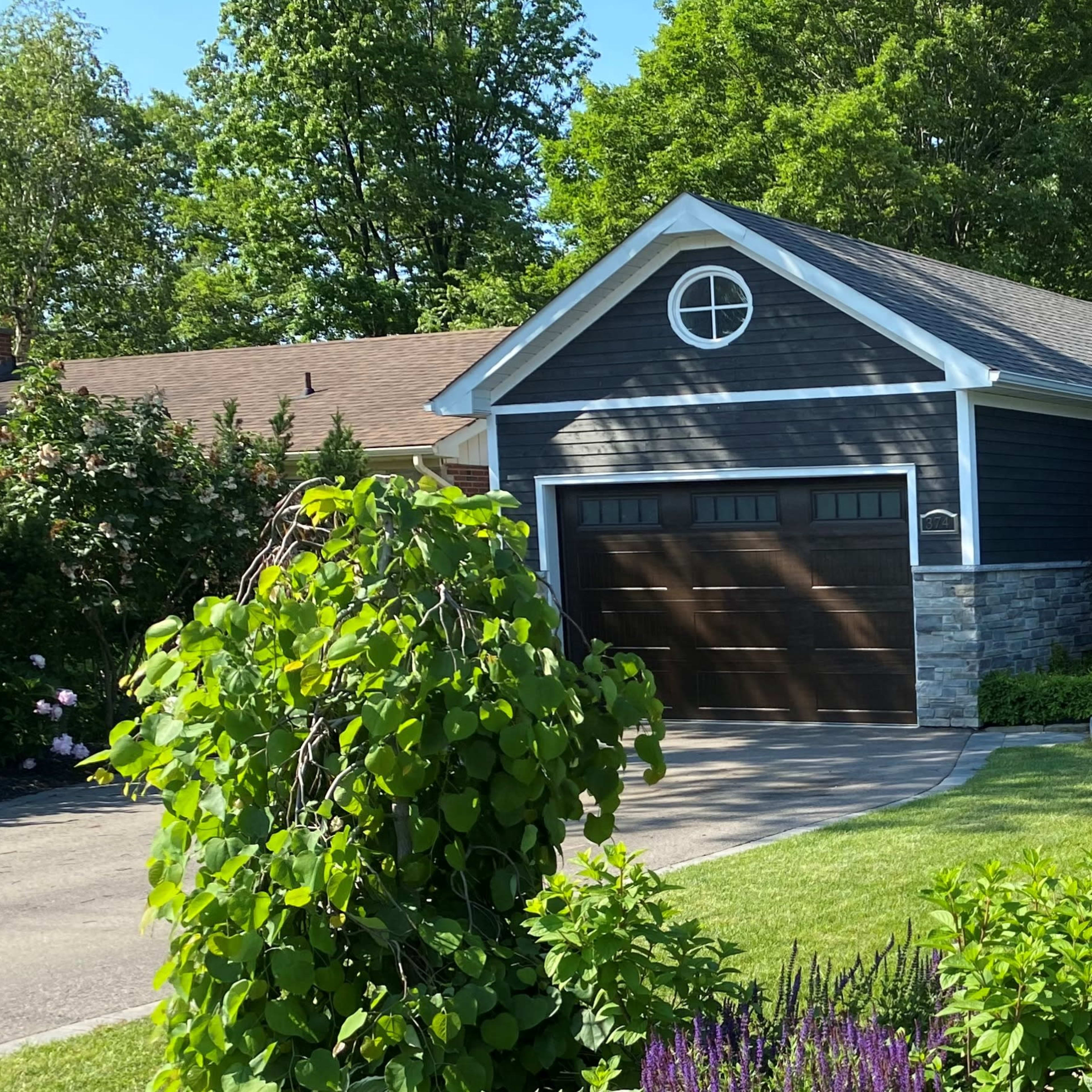 A single-story house with a dark exterior, a stone veneer, and a two-car garage is surrounded by green lawns and landscaping.