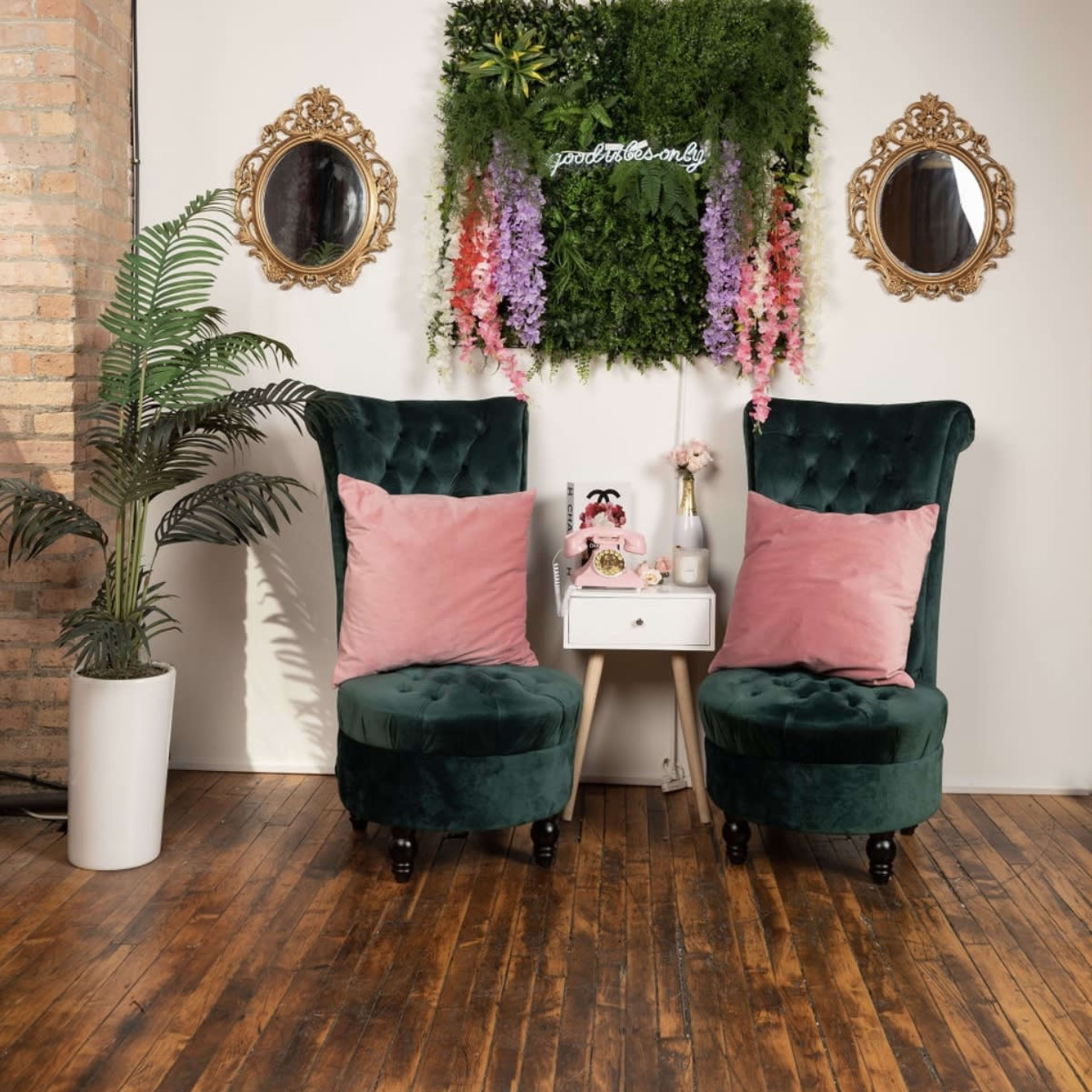Two green velvet chairs with pink throw pillows flanking a small white side table, located in front of a wall adorned with greenery and floral decor, alongside two decorative mirrors.