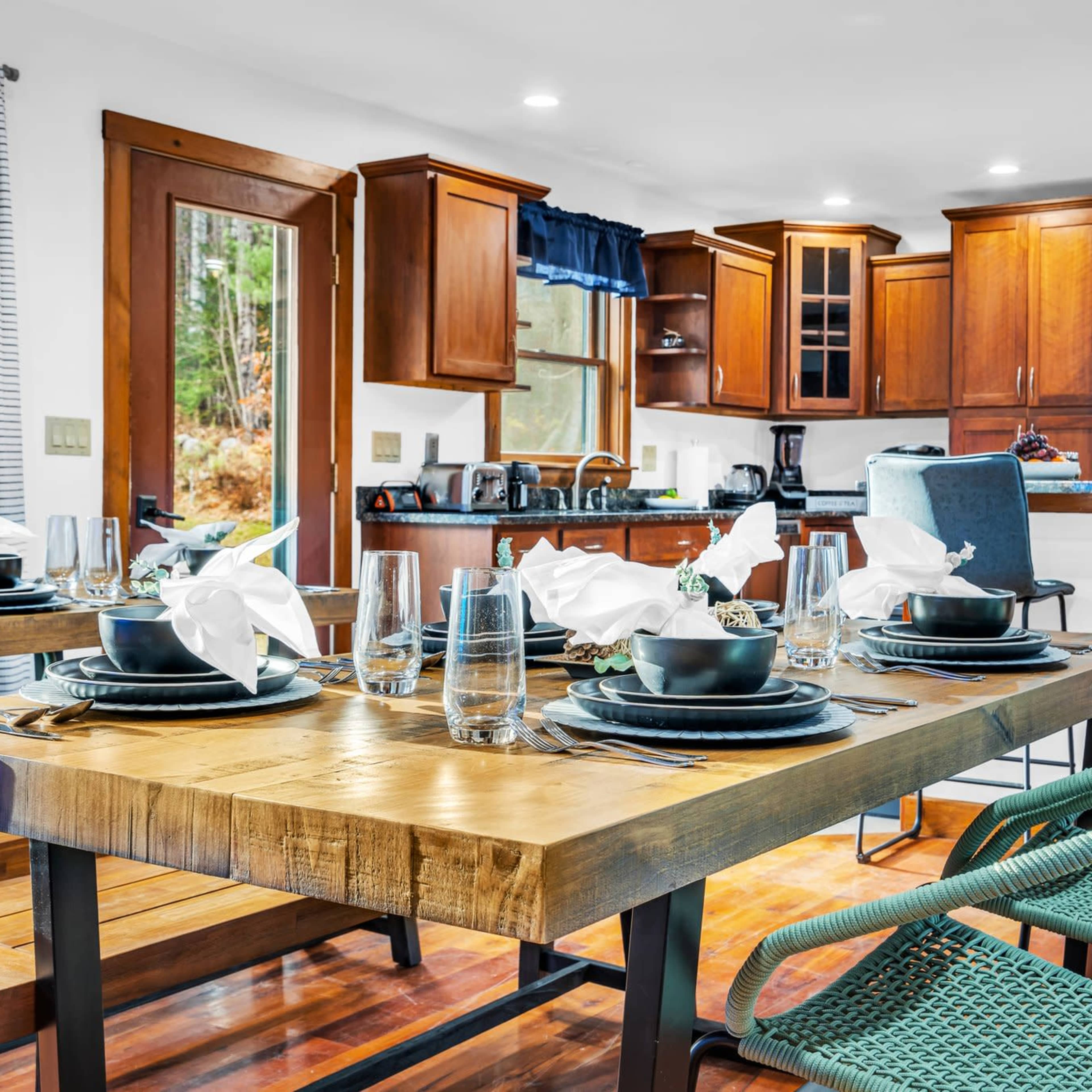 A modern dining area features a wooden table set with dark plates and white napkins, surrounded by turquoise chairs, with an open kitchen in the background.