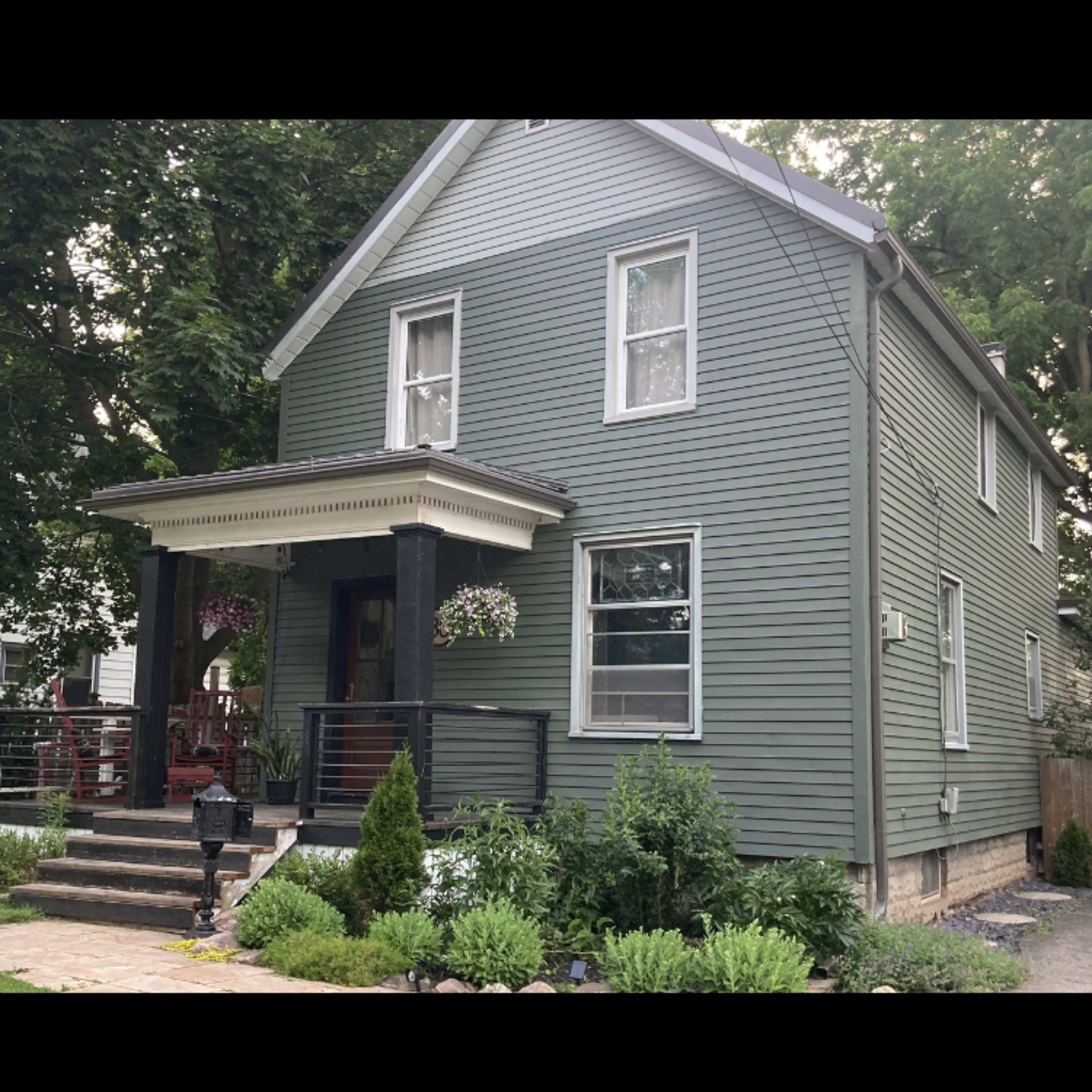 A two-story green house with a front porch, surrounded by shrubs and a landscaped yard.