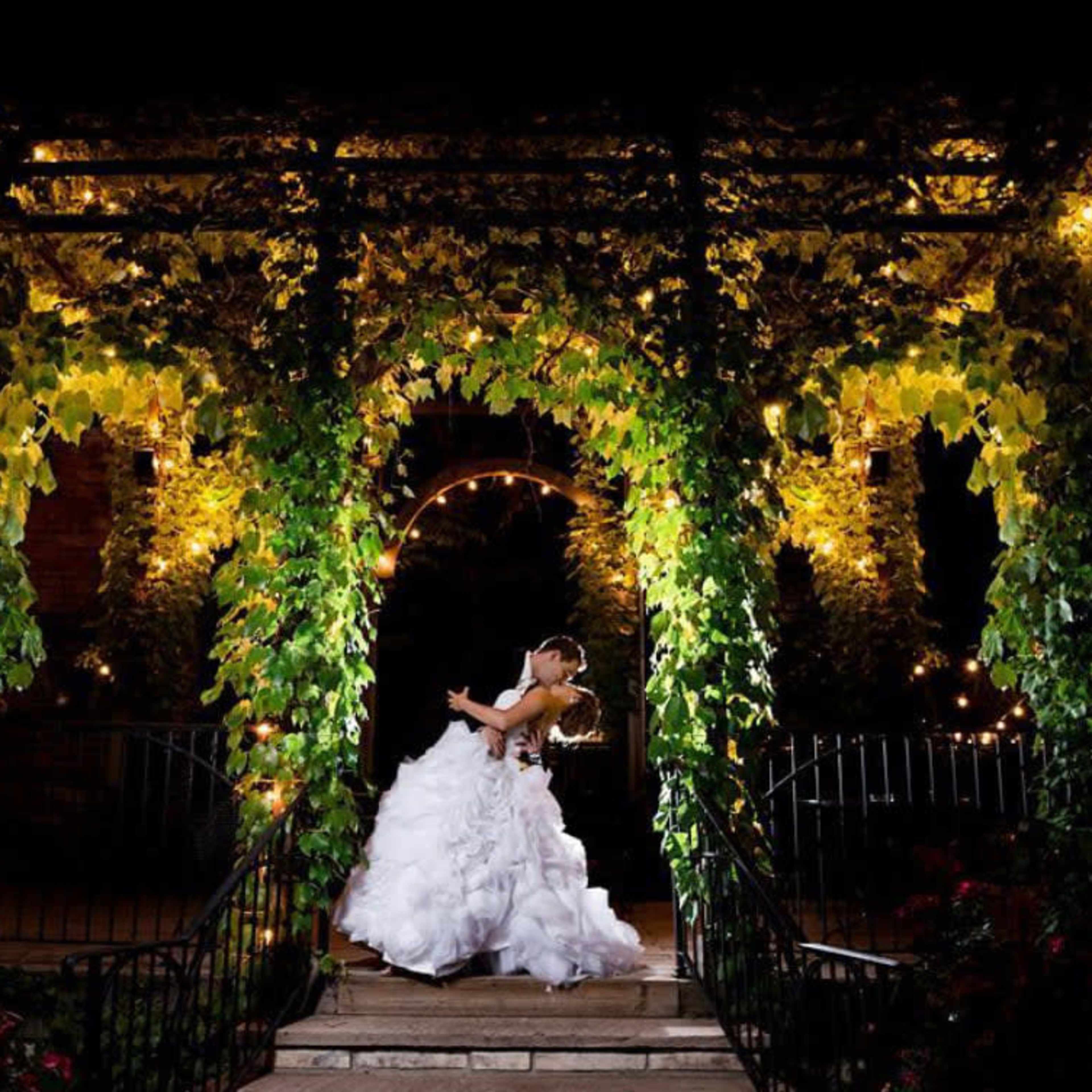 A bride and groom embrace under a vine-covered arch illuminated by warm lights at night.