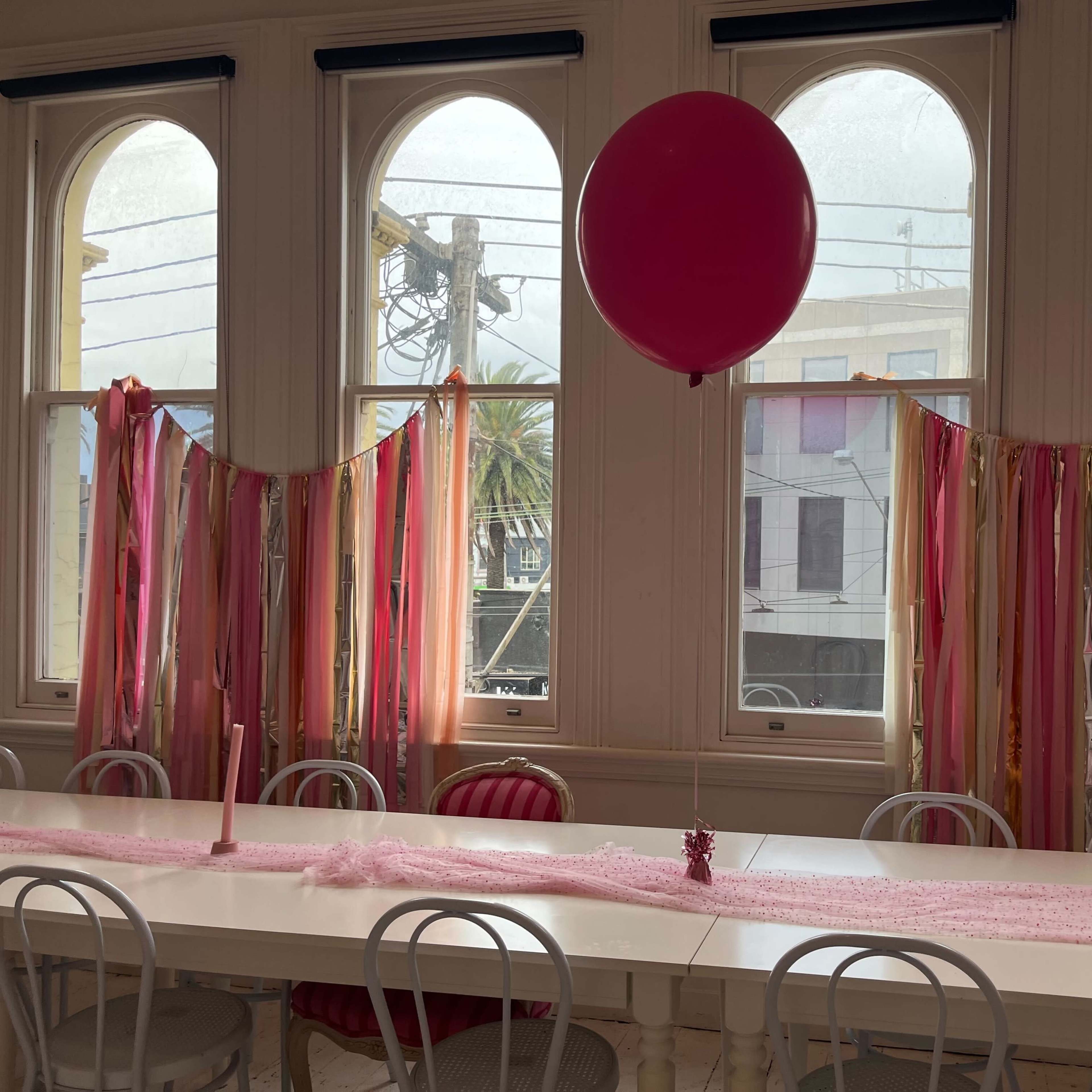 A long table is set up in a brightly lit room with large windows, decorated with pink and gold streamers, candles, and a large pink balloon.