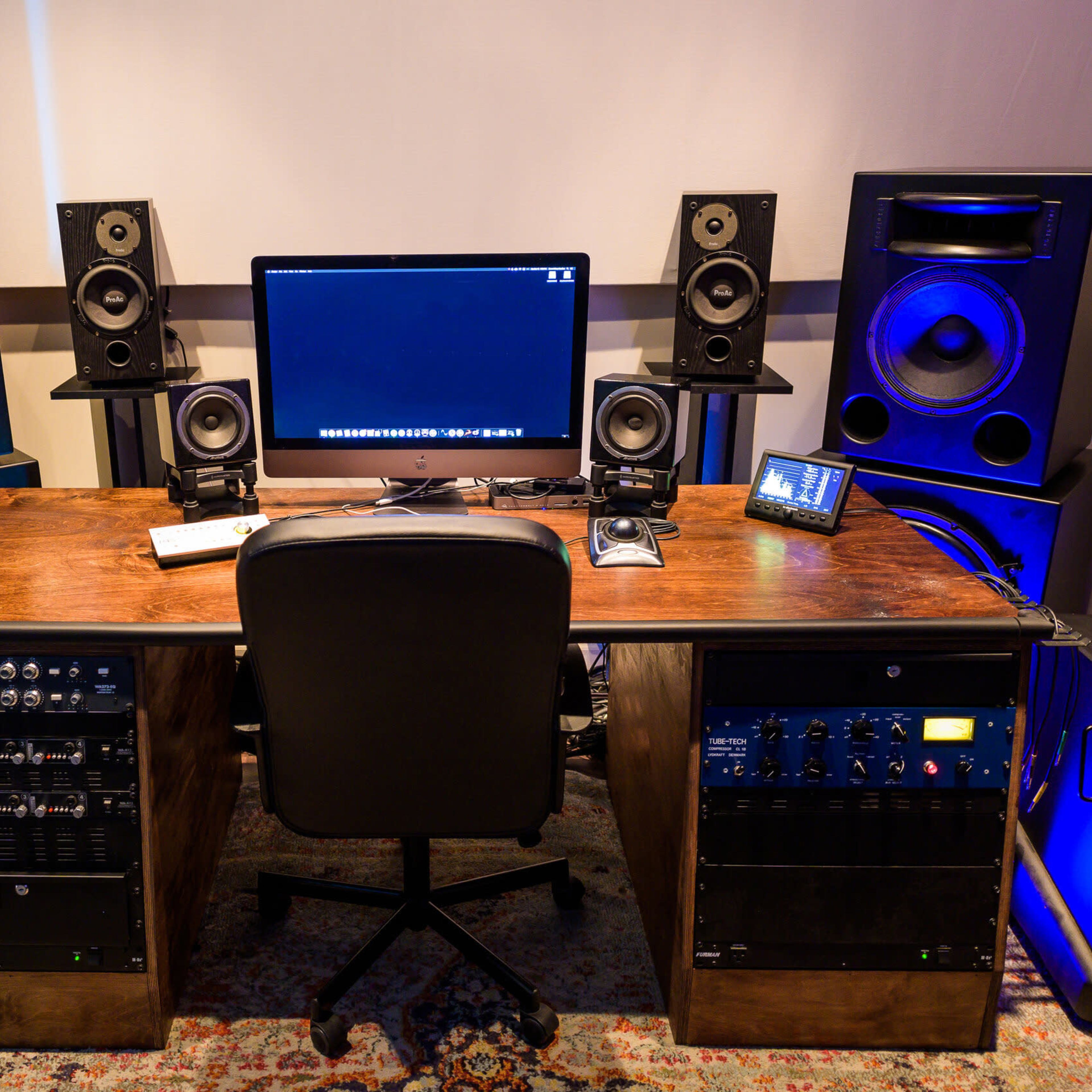A modern audio production studio features a wooden desk with a computer and various music equipment, flanked by large speakers on either side.