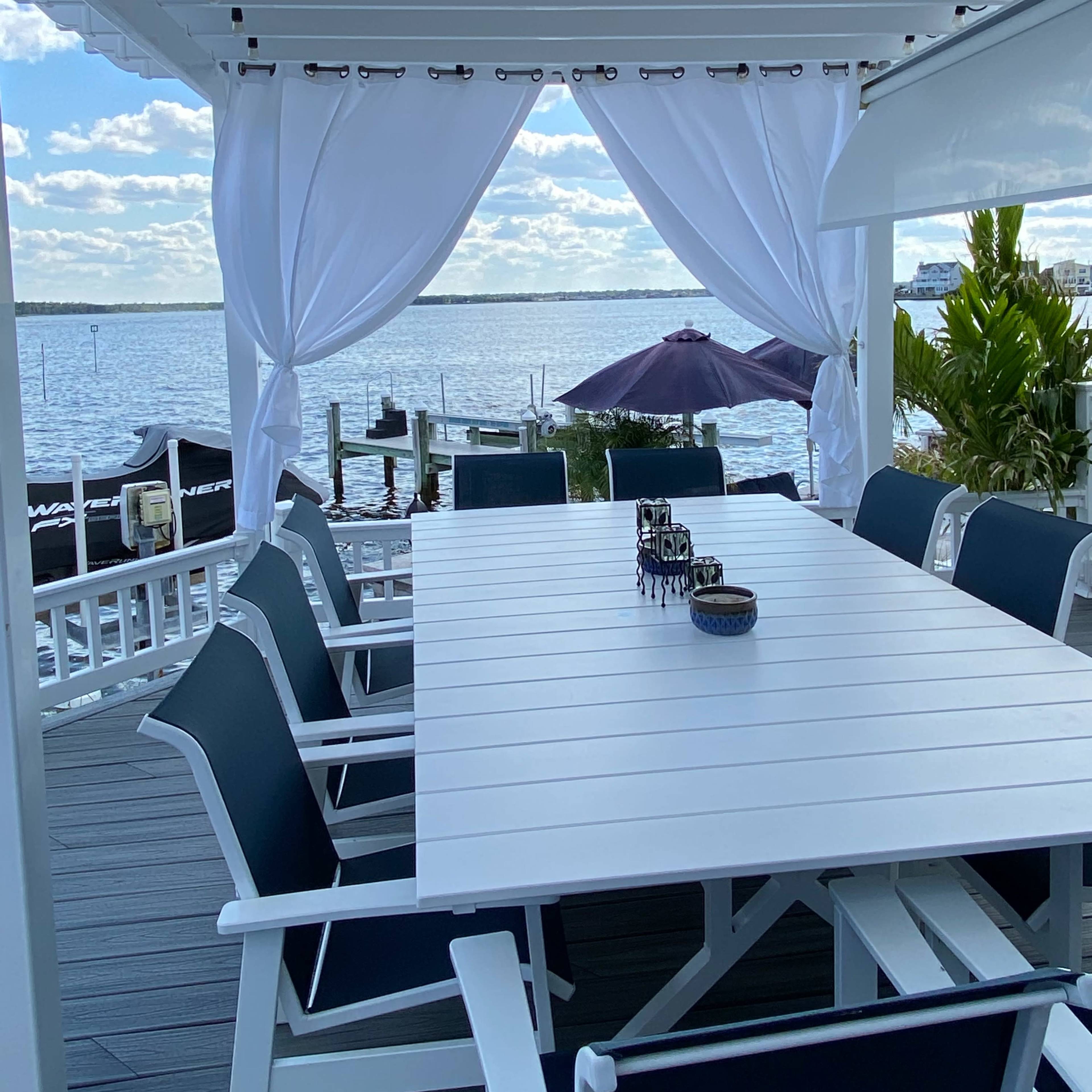 A white table with black chairs is set on a deck overlooking a body of water, framed by flowing white curtains.