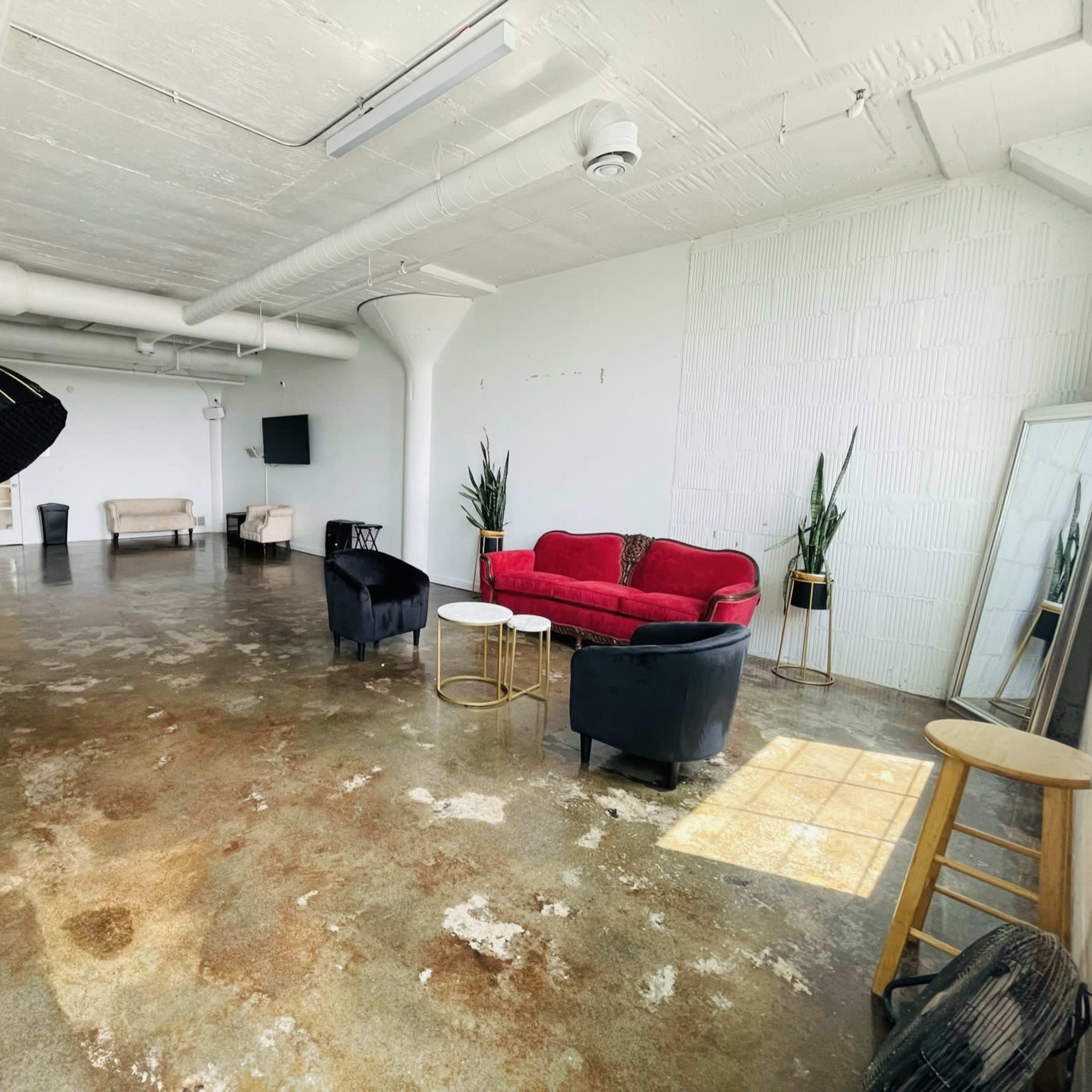 A spacious, empty room with polished concrete floors, featuring a red sofa, two black chairs, and a set of photography equipment.