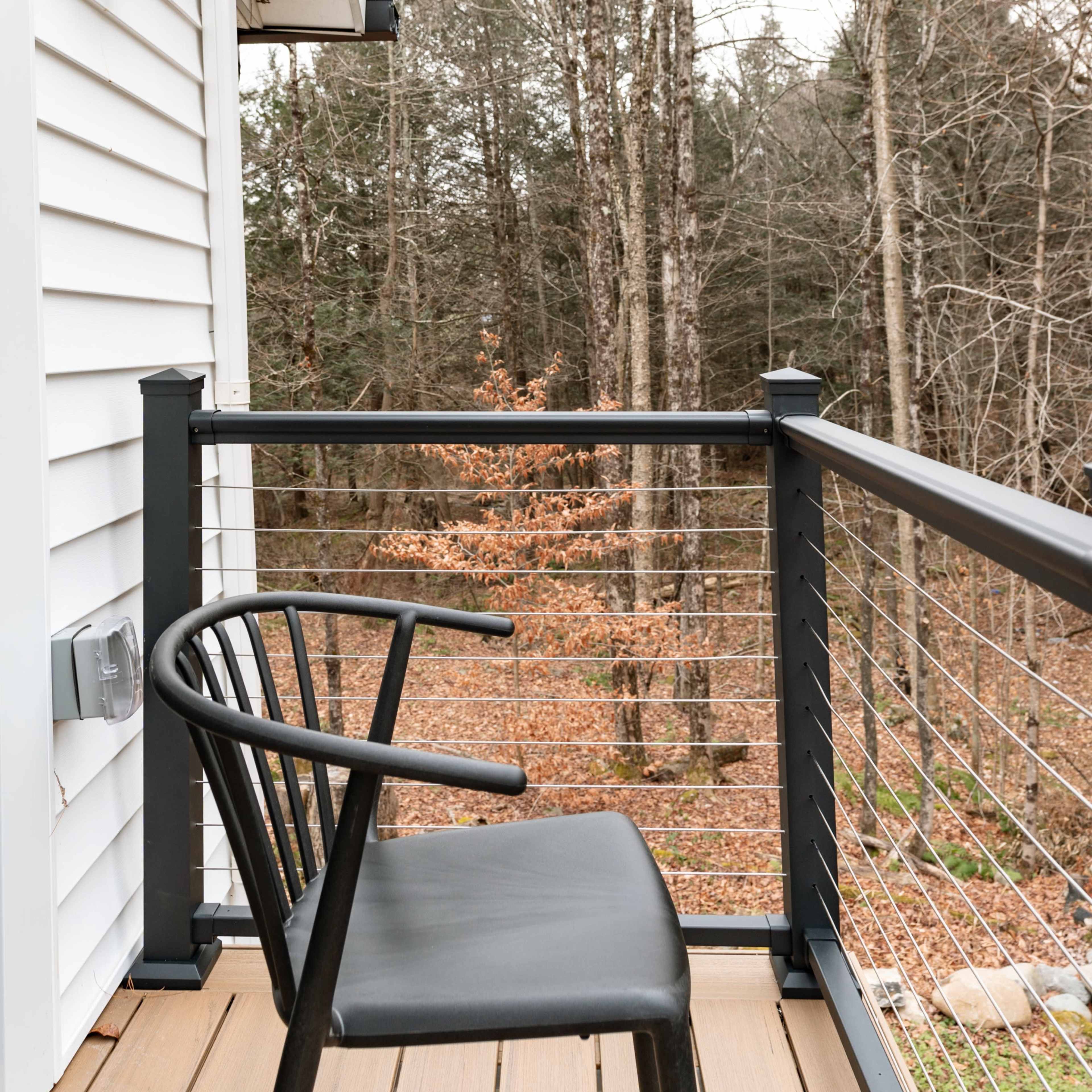 A black chair rests on a wooden balcony overlooking a wooded area with bare trees.