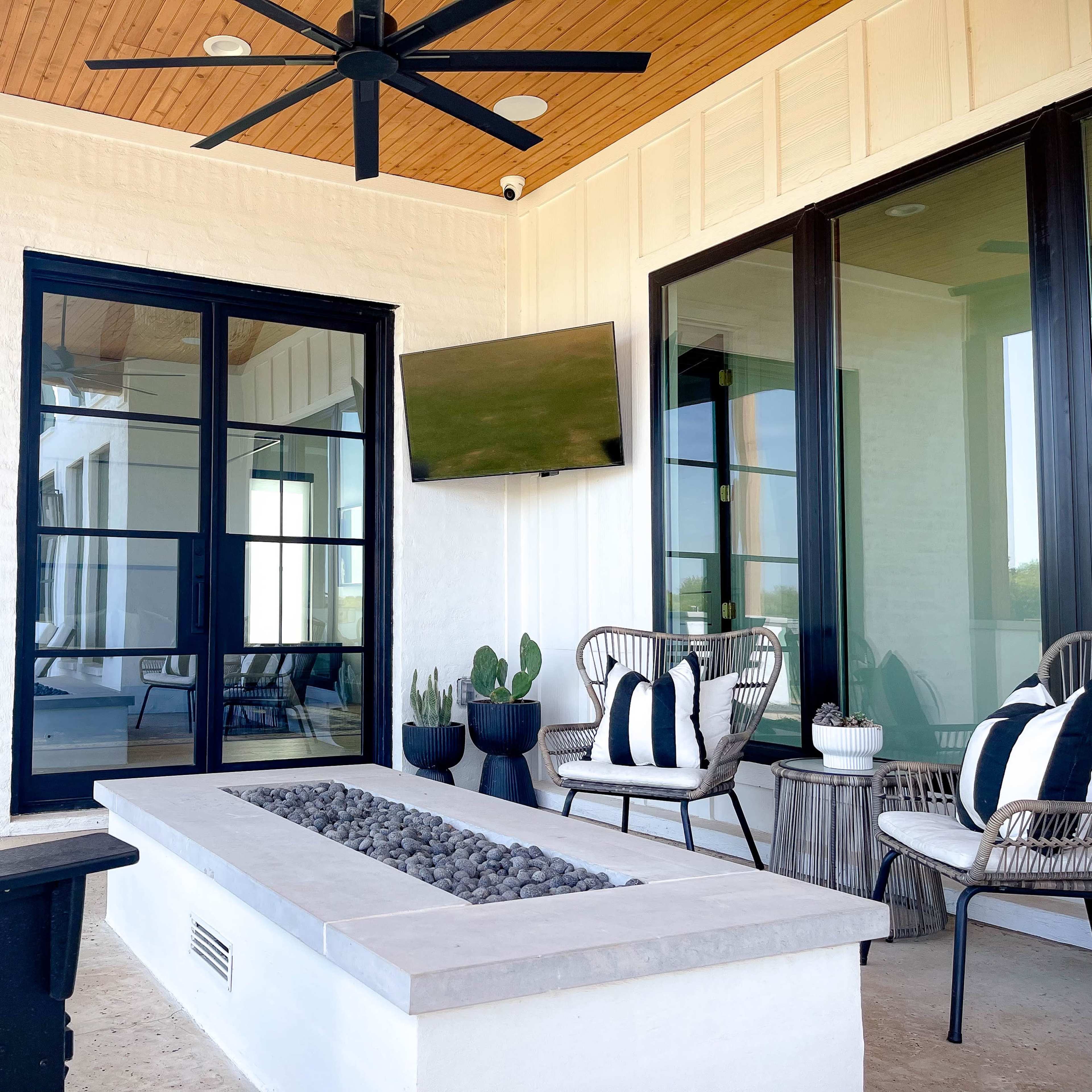 A modern covered patio featuring a seating area with wicker chairs, a fire pit, and a mounted television.