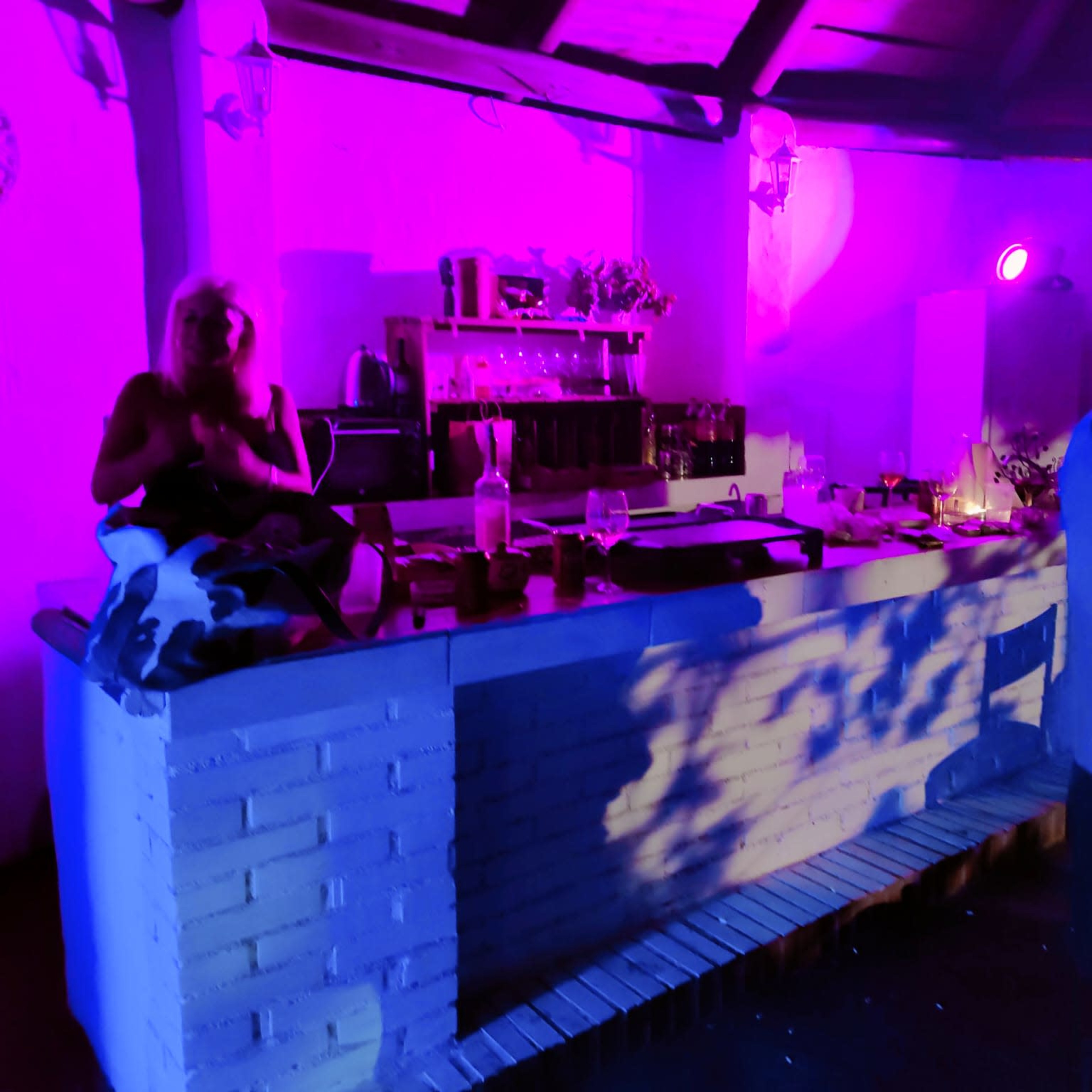A dimly lit kitchen with purple and blue lighting, featuring a counter with various items and two people, one seated and another standing.