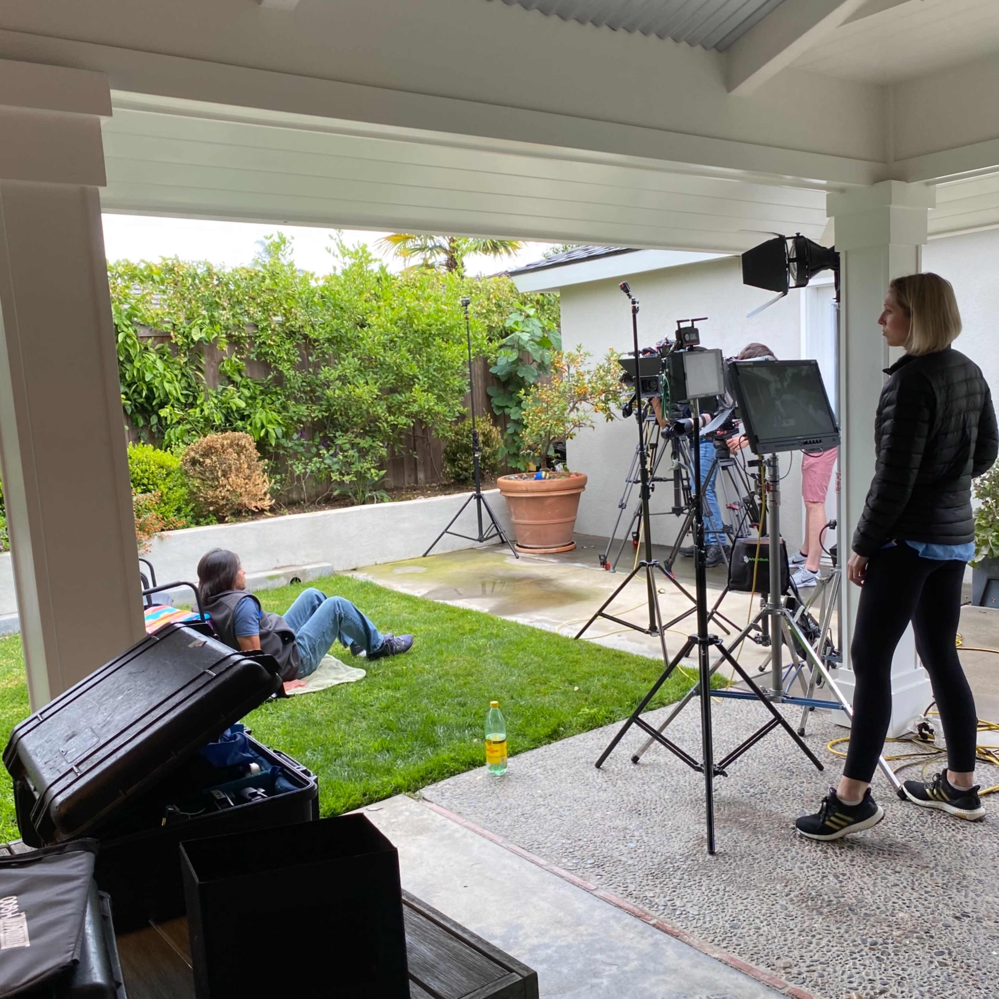 A film crew sets up equipment in a backyard while a person sits on the grass.