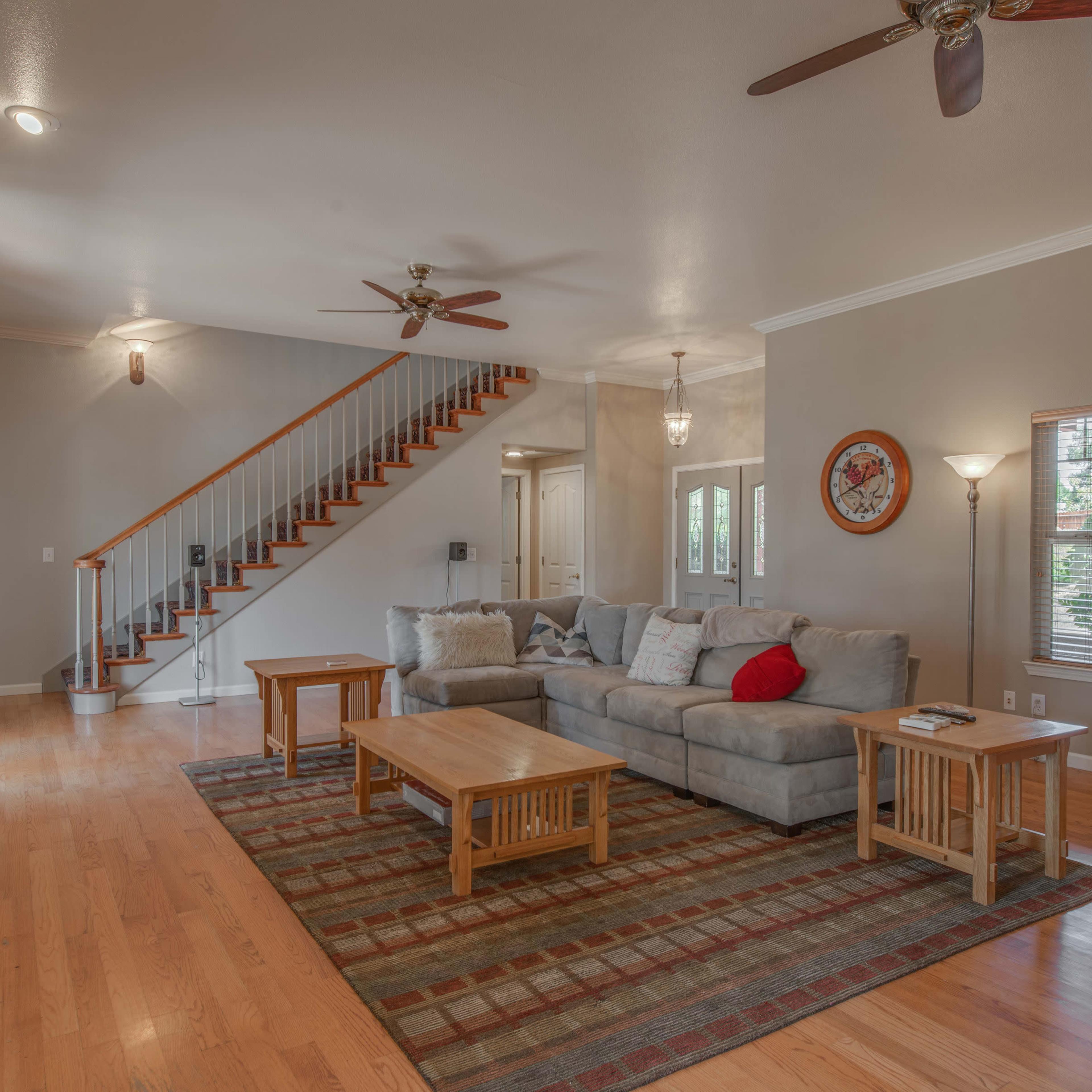 A spacious living room with a stone fireplace, a light gray couch, a wooden coffee table, and large windows, featuring a staircase in the background.
