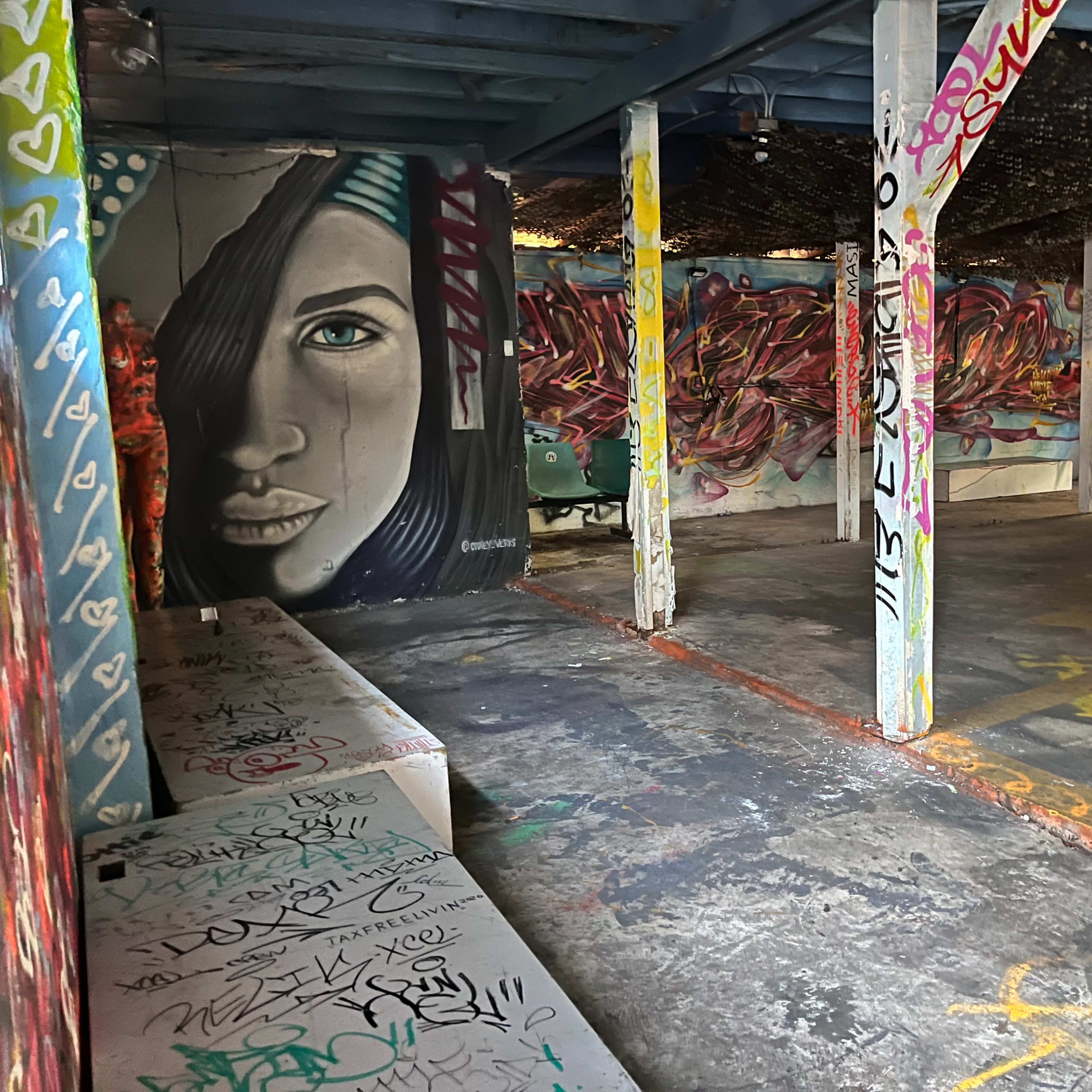 An indoor space with graffiti-covered walls and a large mural of a face, surrounded by colorful tags and a few tables set up in the area.