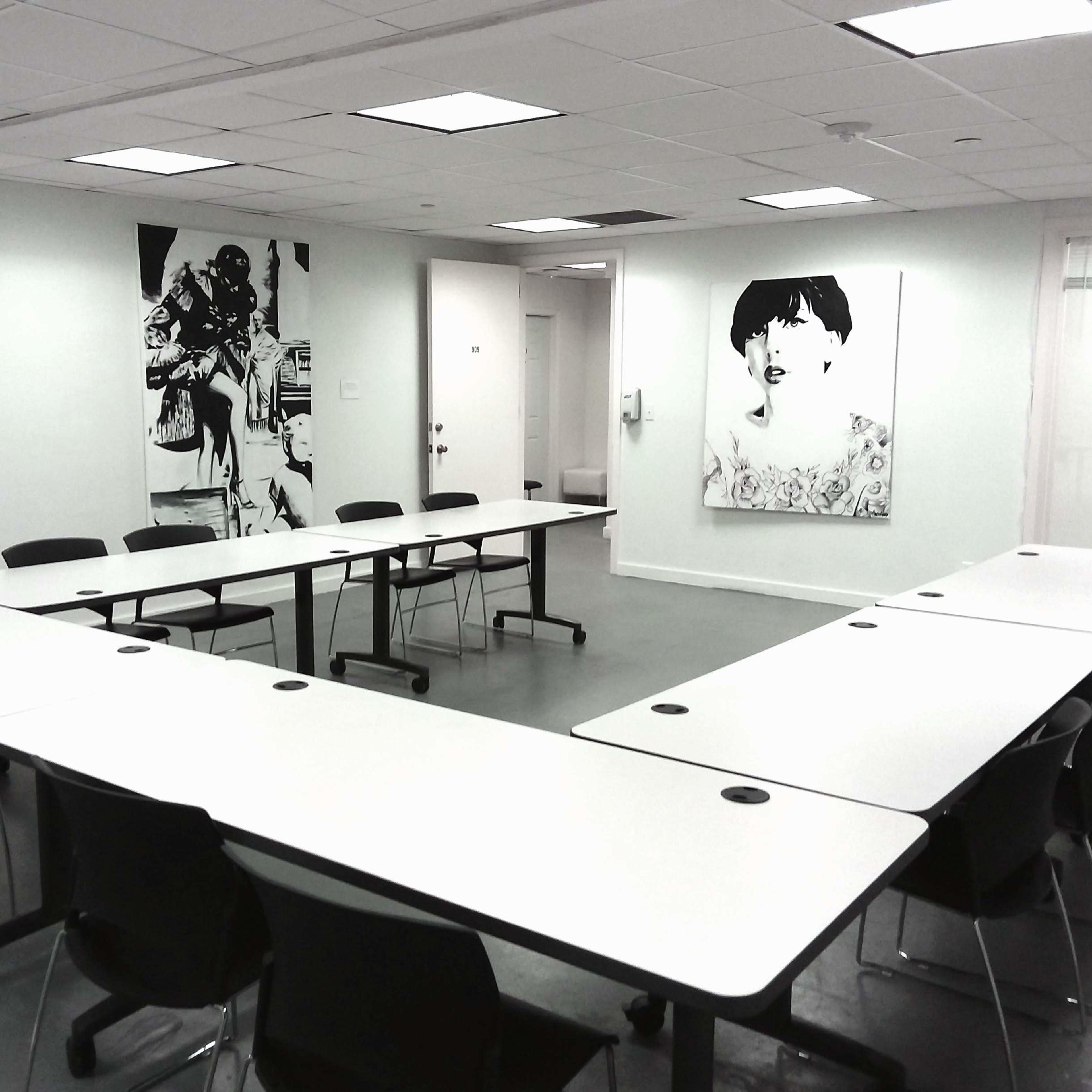 A conference room features a U-shaped arrangement of white tables and black chairs, with large framed artworks on the walls.