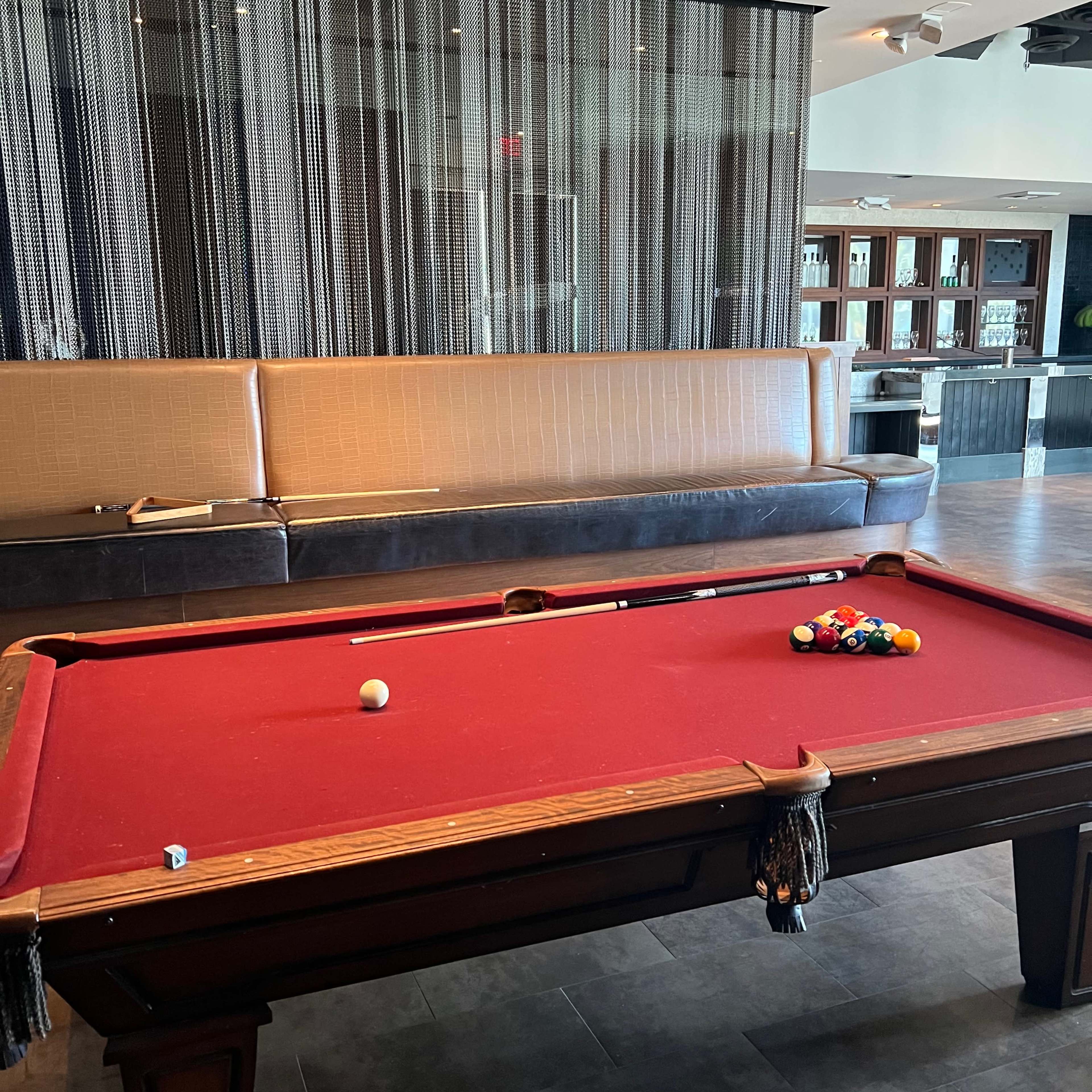 A pool table with a red felt surface and a triangle of colored balls sits in a lounge area with a brown leather bench and a modern design.