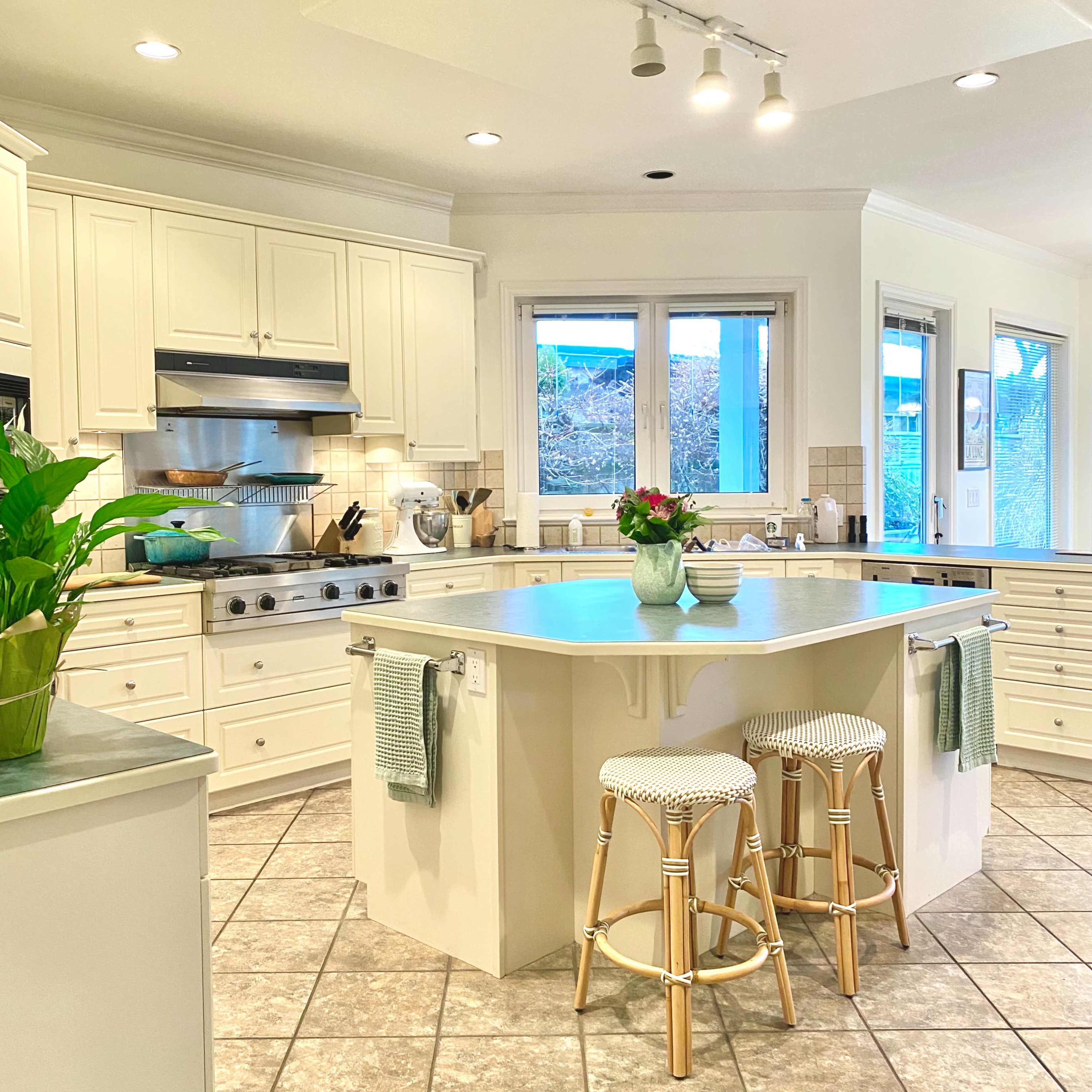 A modern kitchen features light-colored cabinetry, a central island with two stools, and a large window overlooking a garden.
