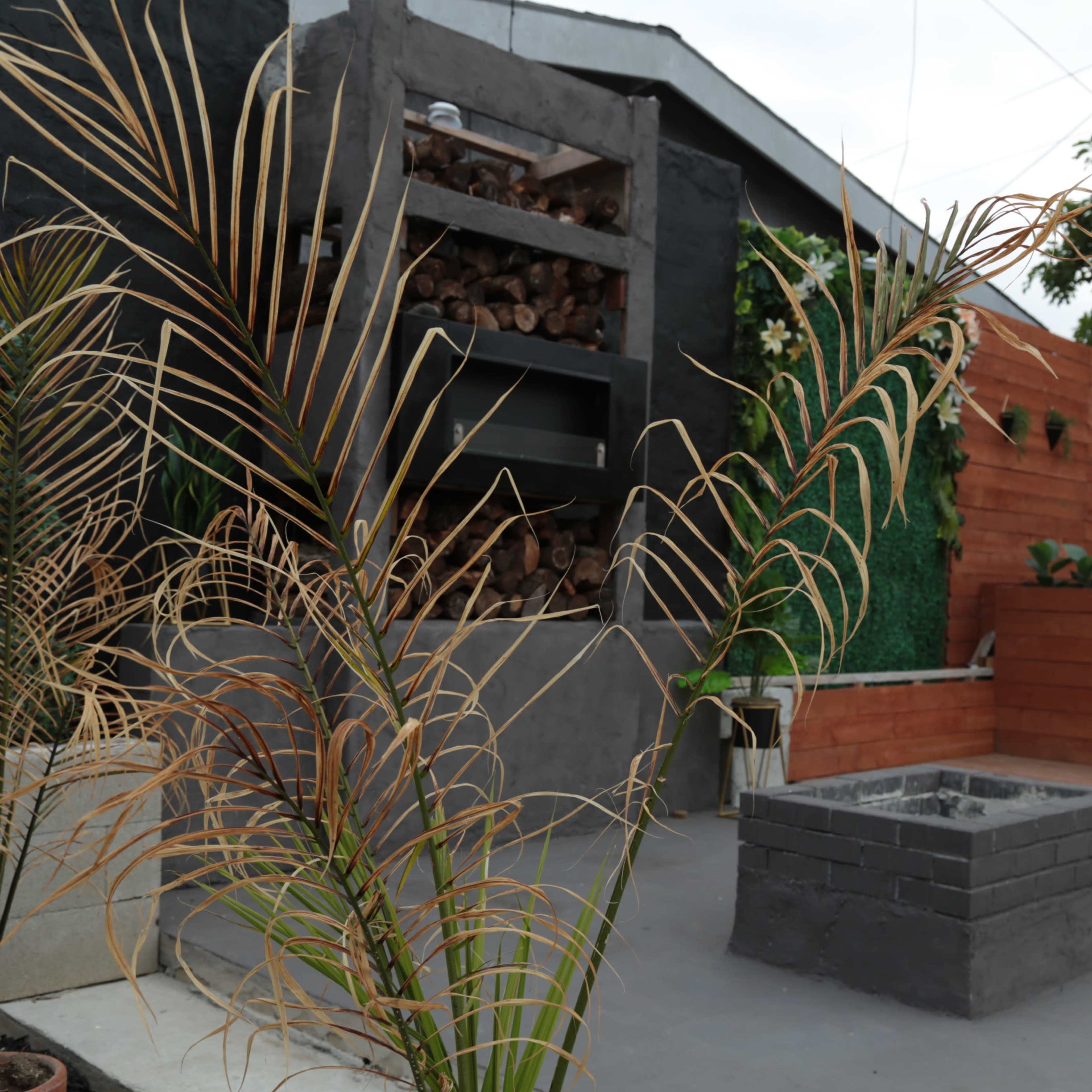 A modern outdoor patio area featuring a stone fire pit, vertical wood storage, and landscaped greenery.
