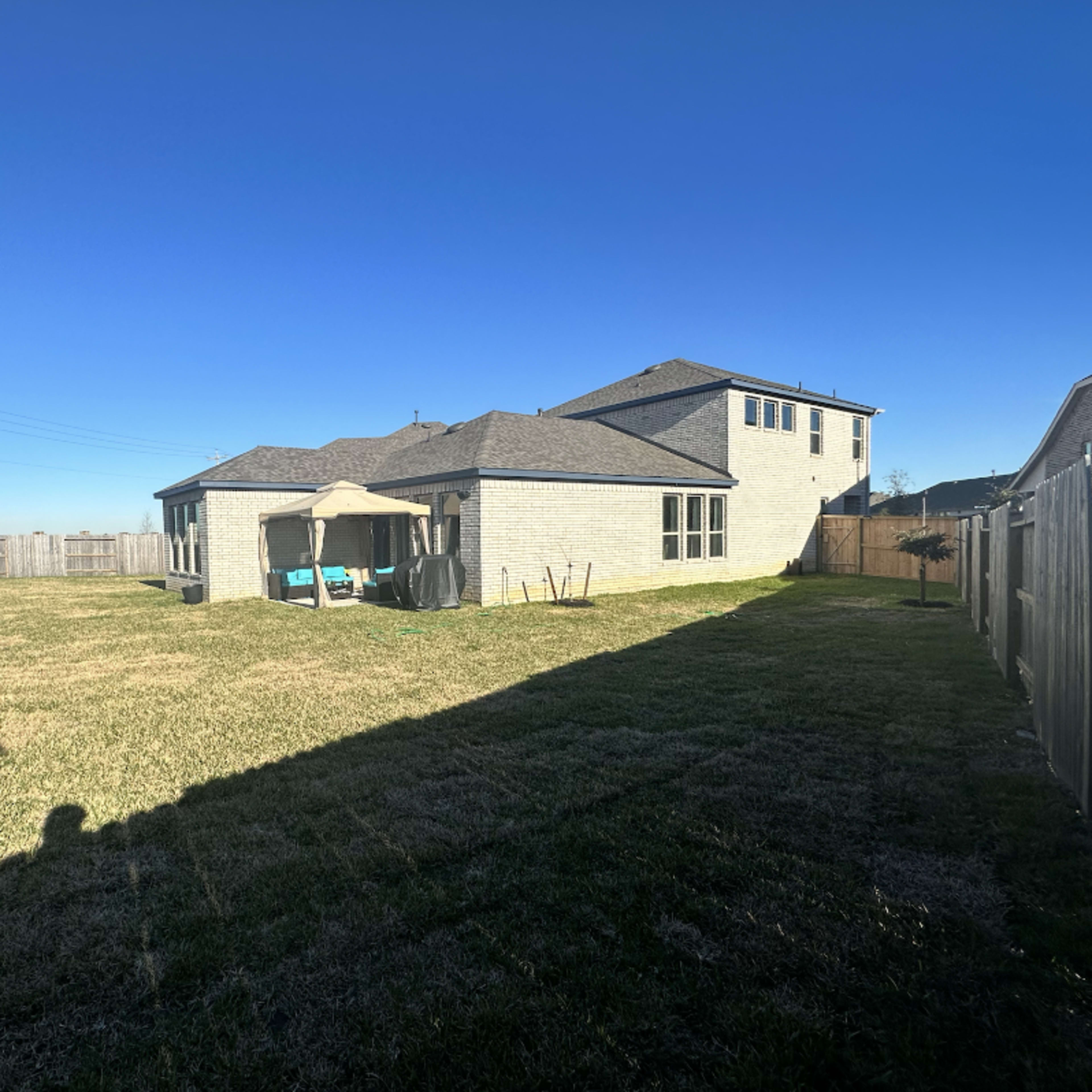 A spacious backyard features a house with a gray exterior, a patio canopy, and an expansive lawn surrounded by a fence.