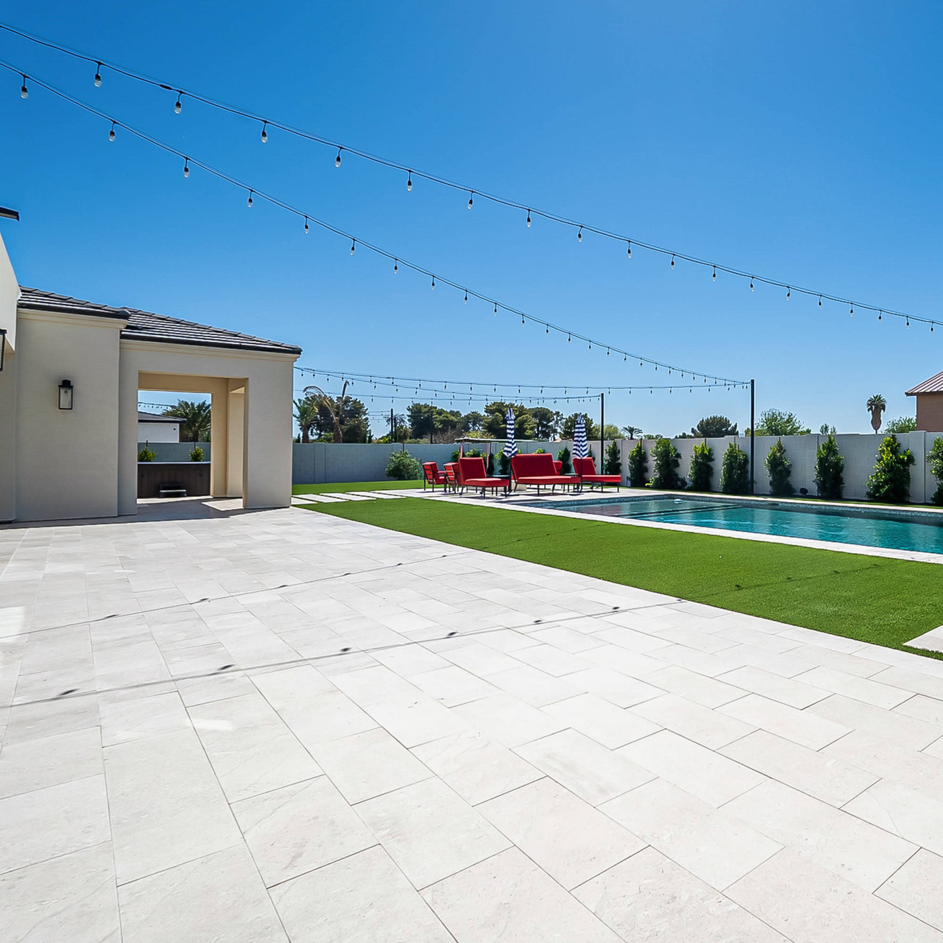 A modern backyard featuring a swimming pool, surrounded by stone tiles and artificial grass, with red outdoor furniture and string lights overhead.