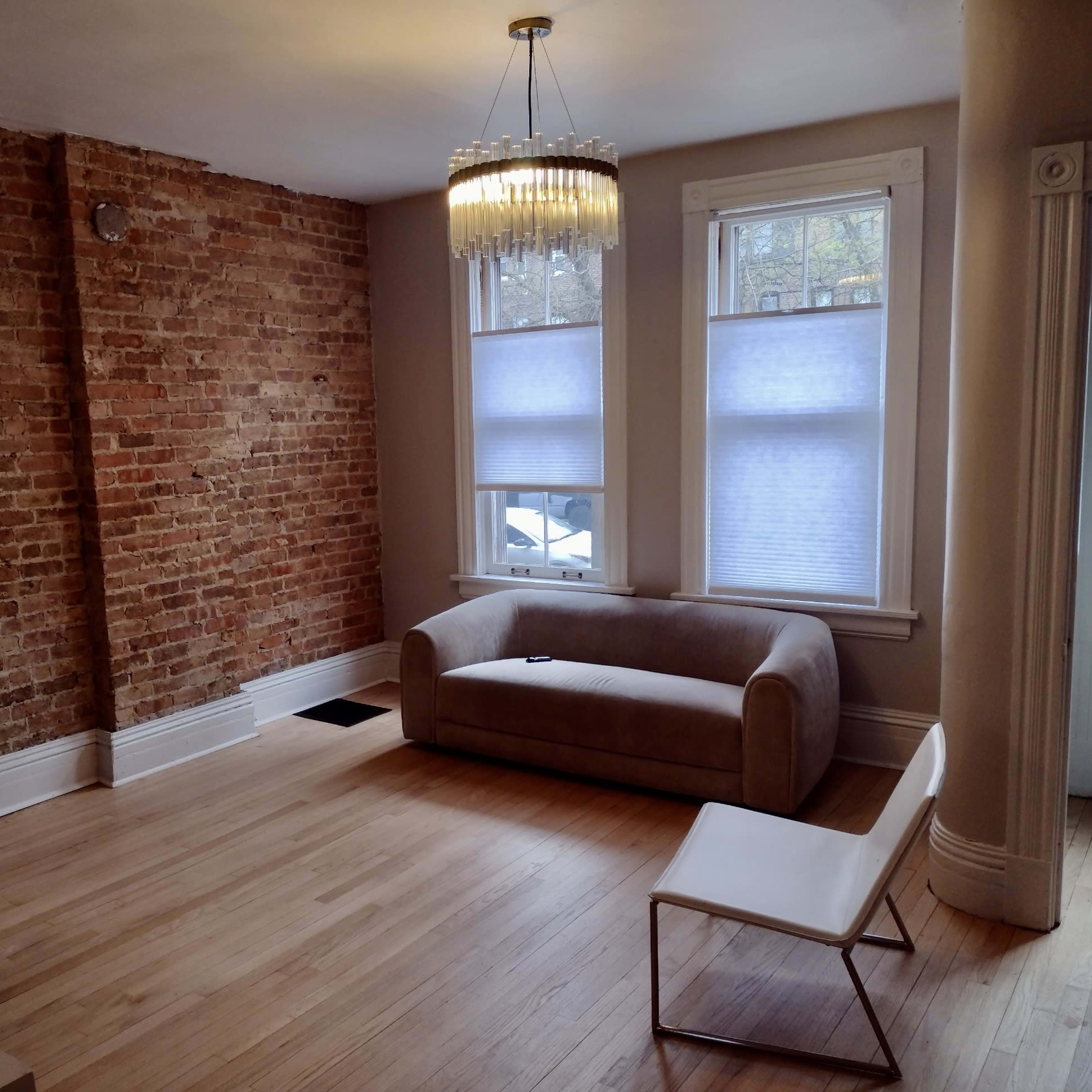 A living room with exposed brick walls, a light-colored sofa, a modern chair, and large windows with shades.