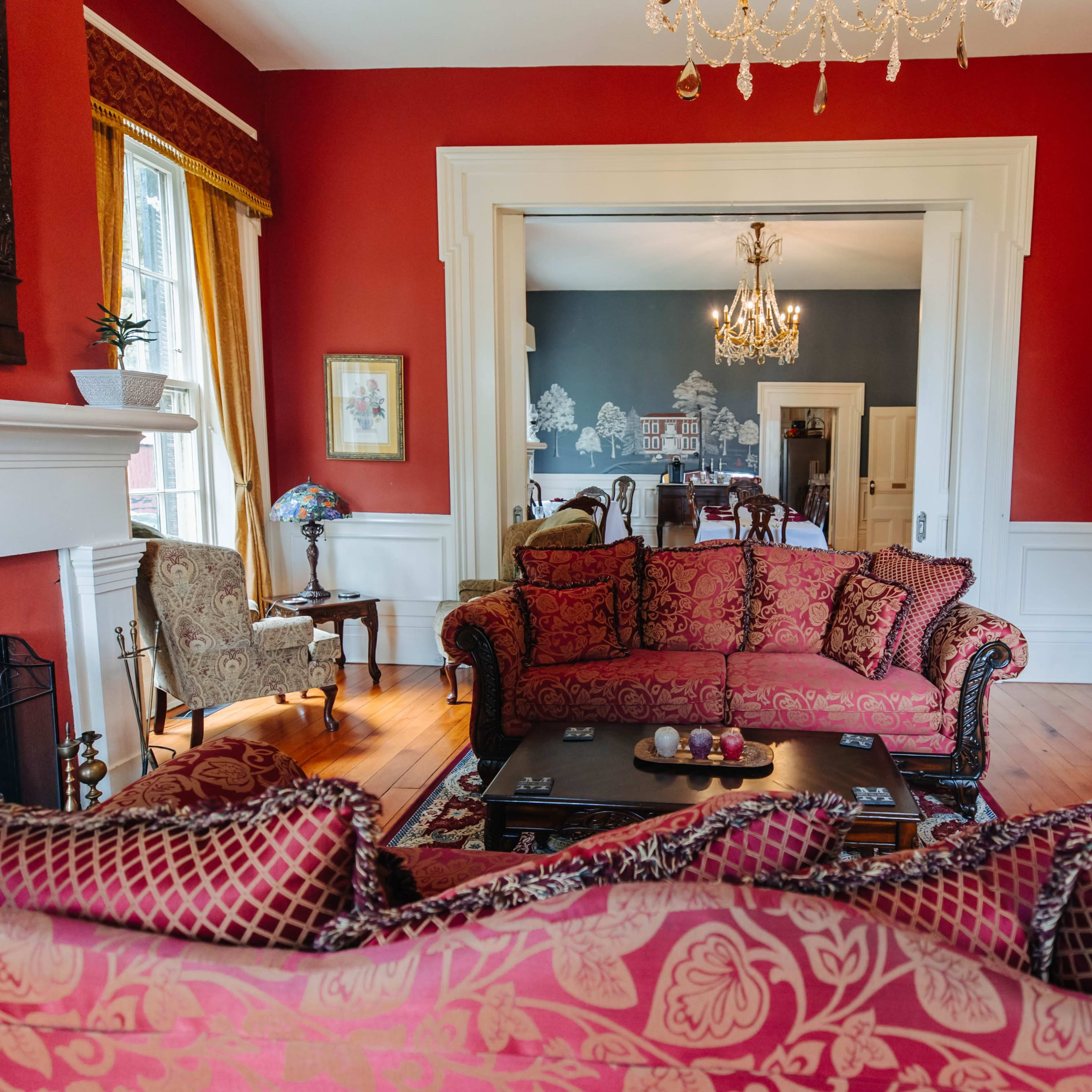 A richly decorated living room featuring a deep red wall, a patterned sofa, and a view into a dining area with a chandelier.