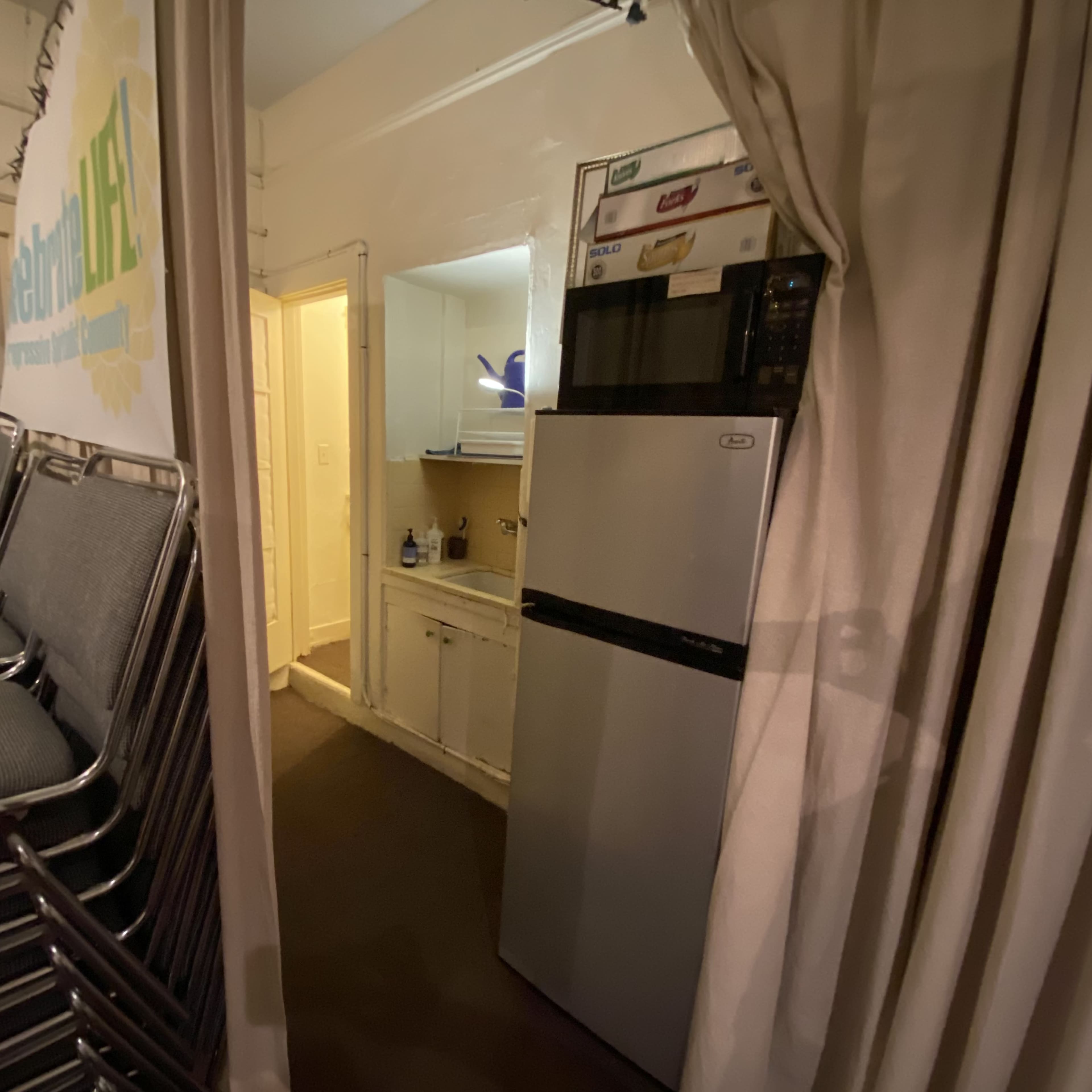 A narrow hallway leading to a kitchen area with a refrigerator and a doorway beyond it, next to a stack of metal chairs.