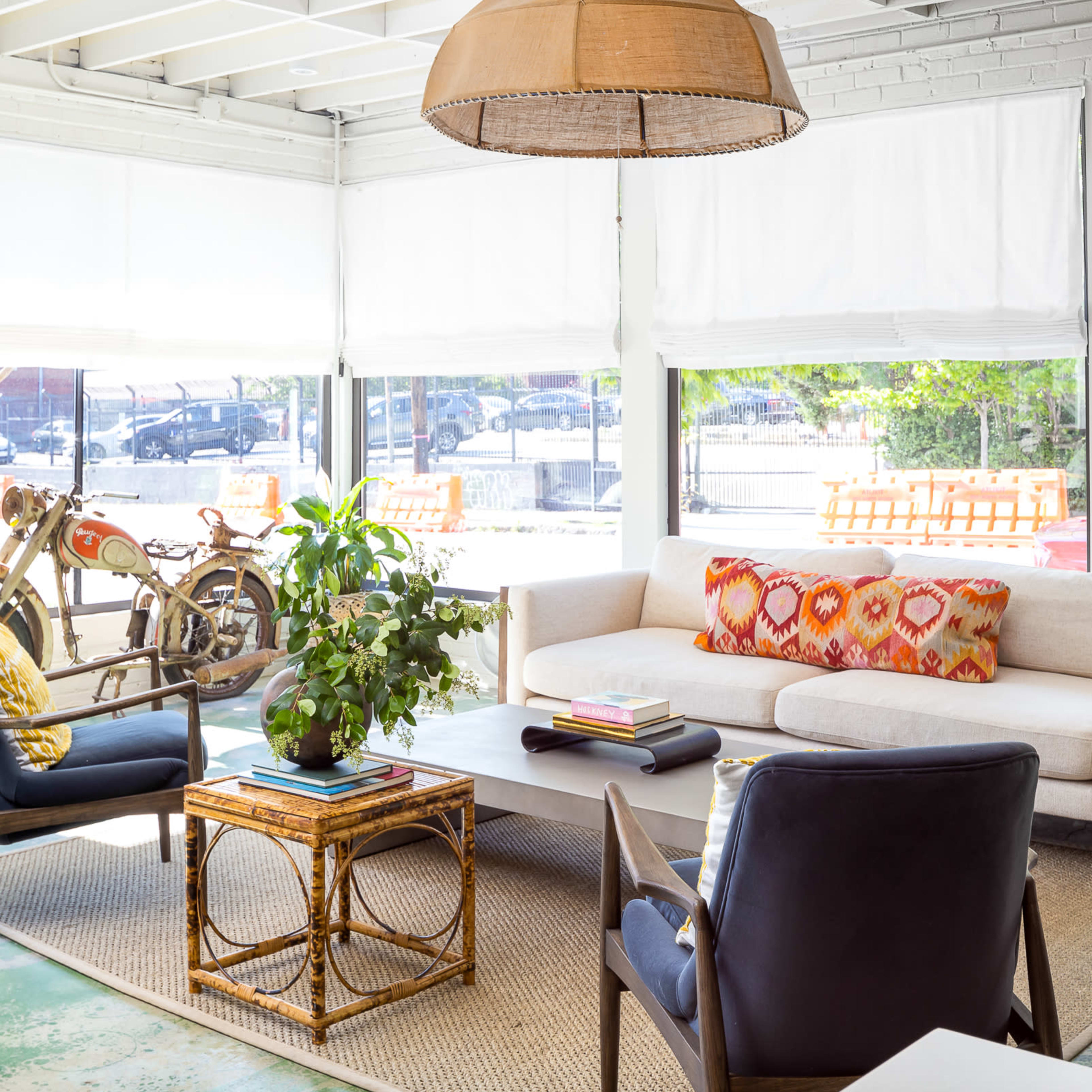A bright living space featuring a beige sofa with a colorful cushion, a coffee table, a potted plant, and large windows with natural light, alongside a bicycle and a parked car visible outside.