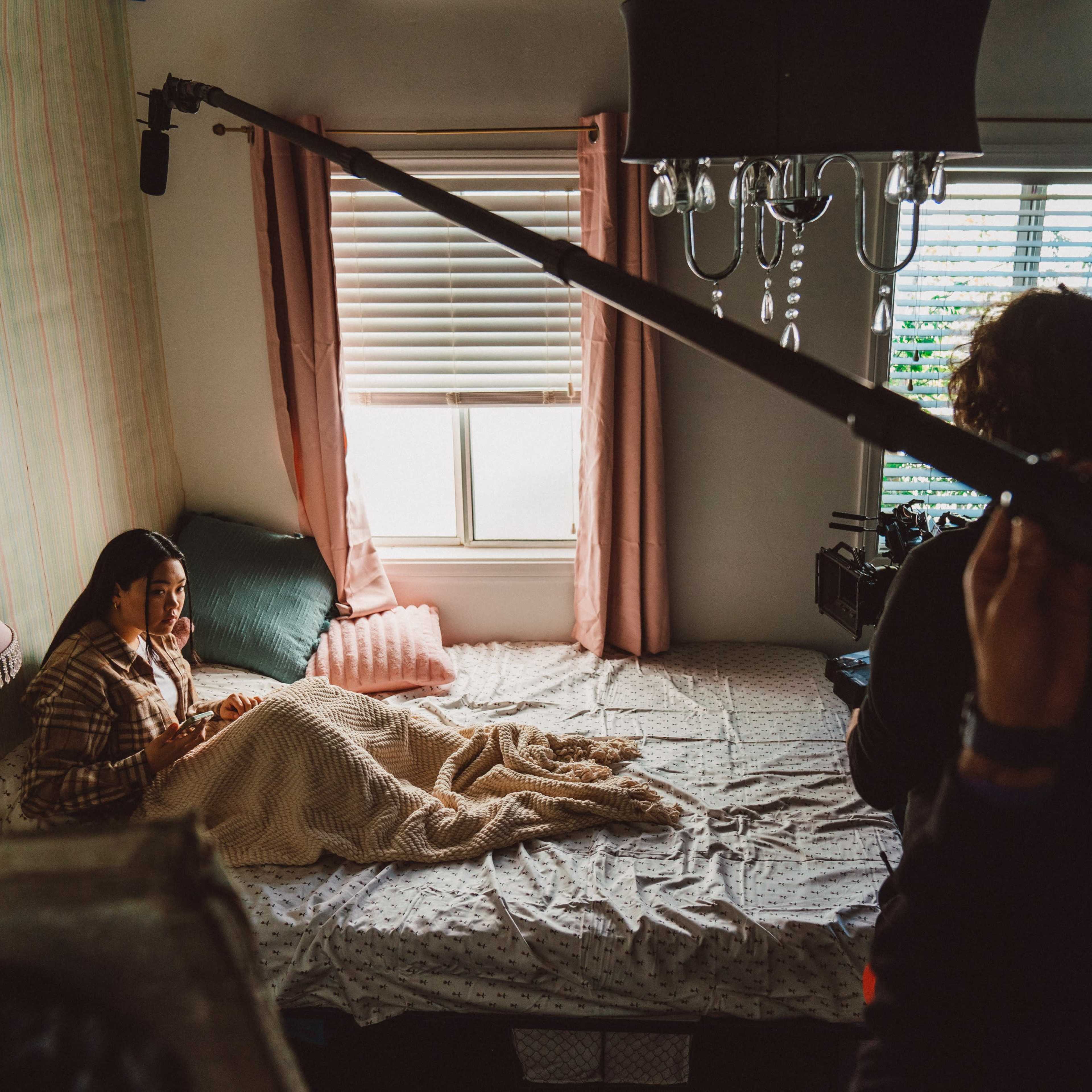 A woman sits on a bed in a softly lit room while a crew member adjusts a microphone positioned above her.
