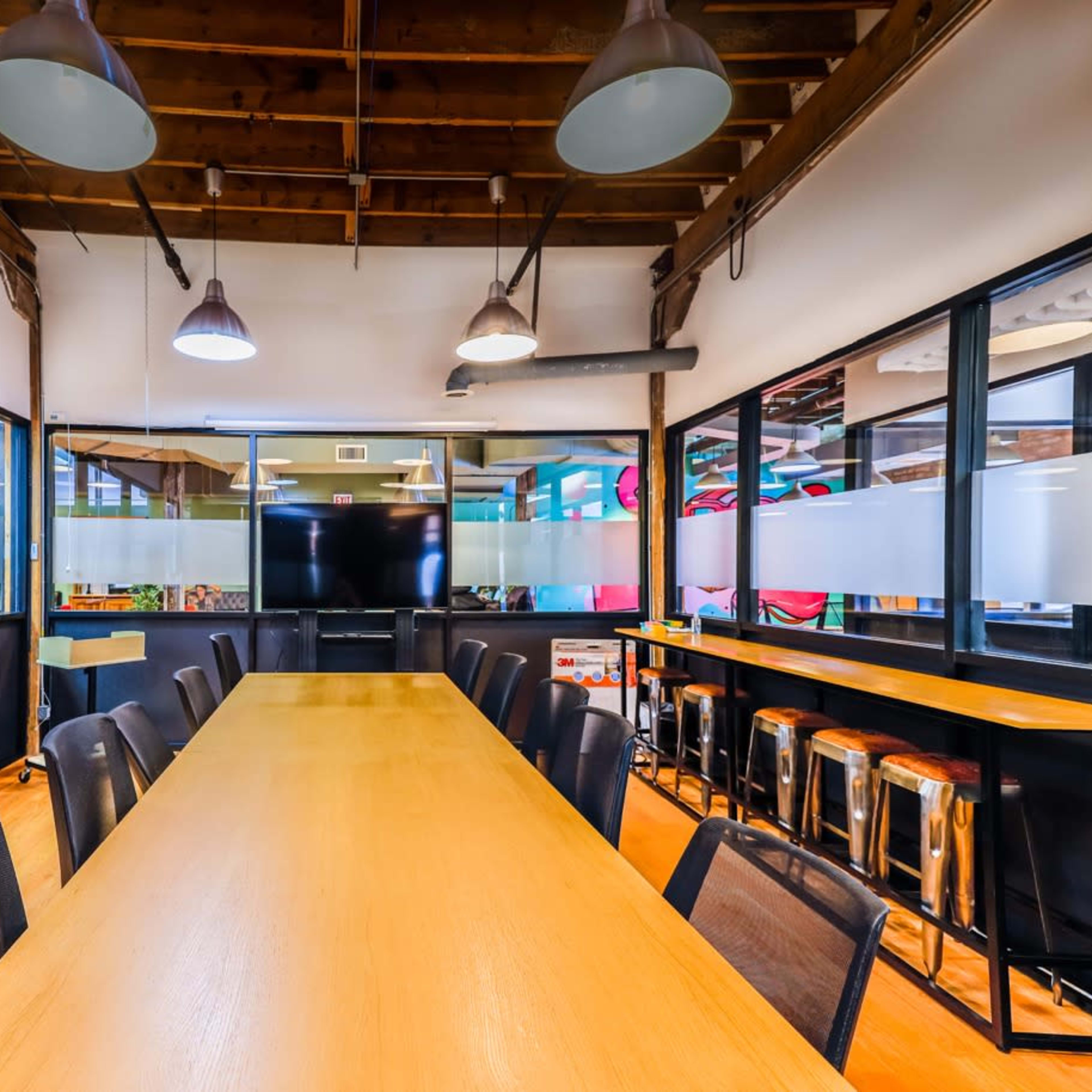 A long wooden conference table is situated in a room with modern lighting and large windows, featuring a wall-mounted screen at one end.