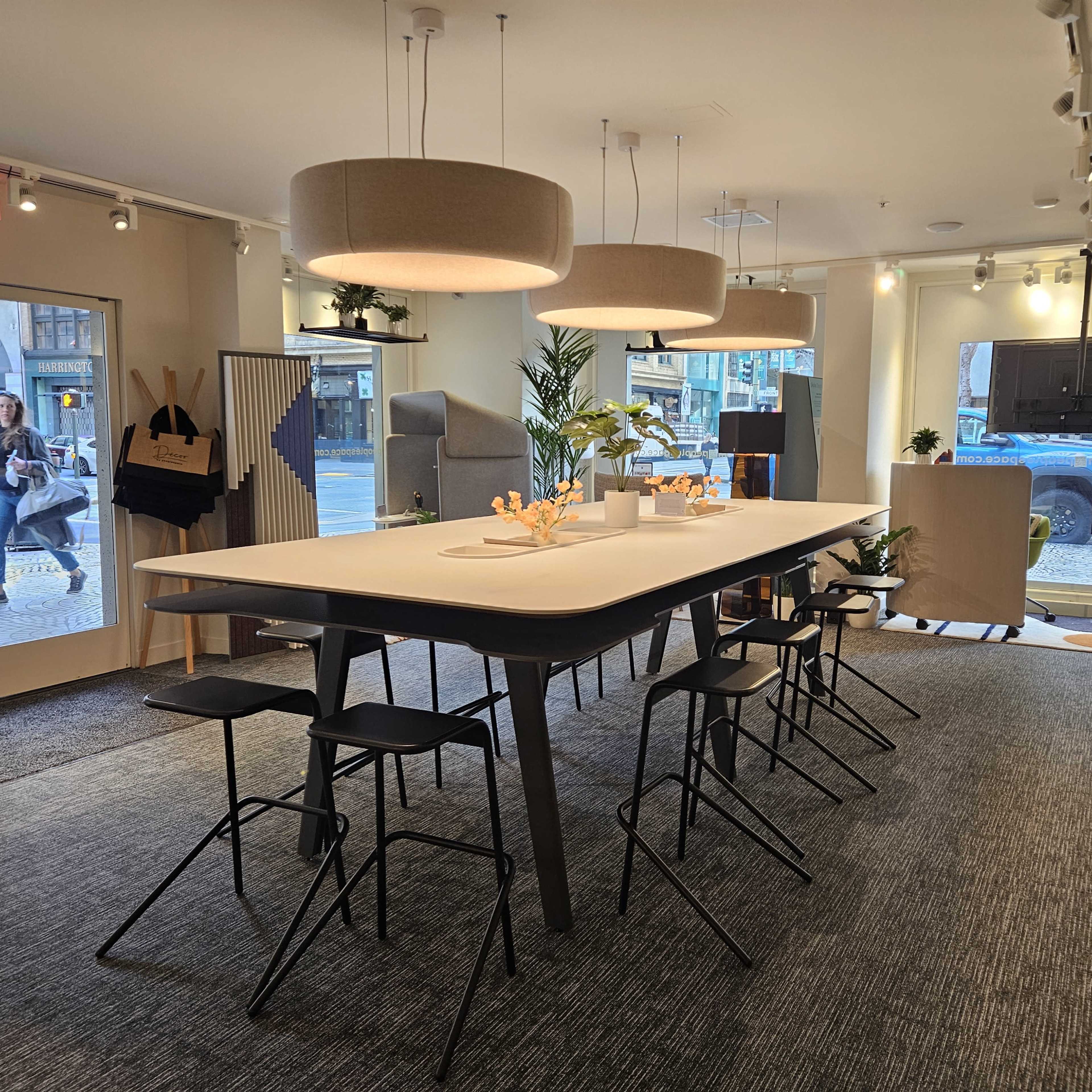 A modern interior space with a large table surrounded by high stools, well-lit by pendant lights, and featuring a large window displaying a street view.